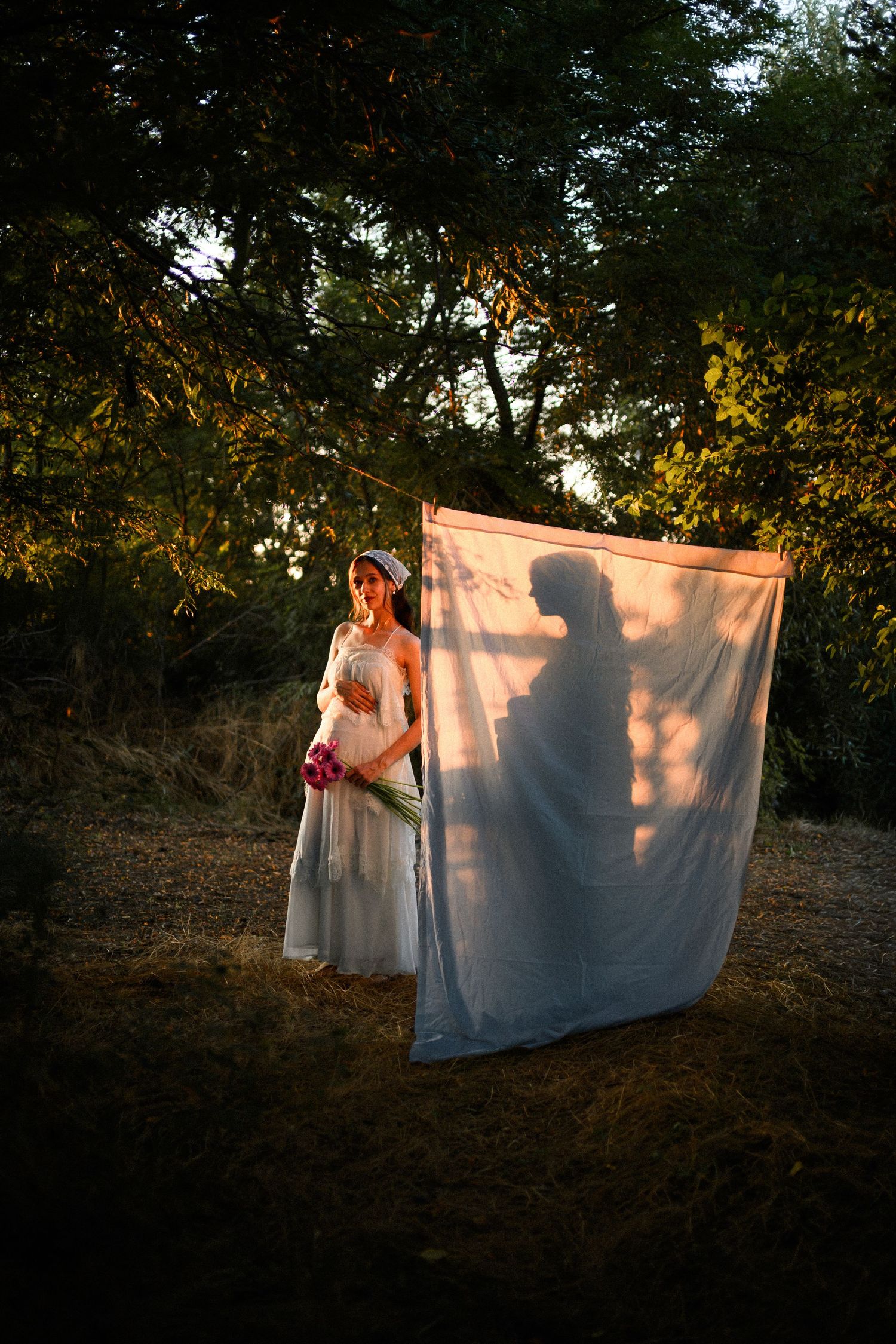Bride in white dress holds flowers outdoors, with a dramatic shadow of a couple projected on a white sheet.