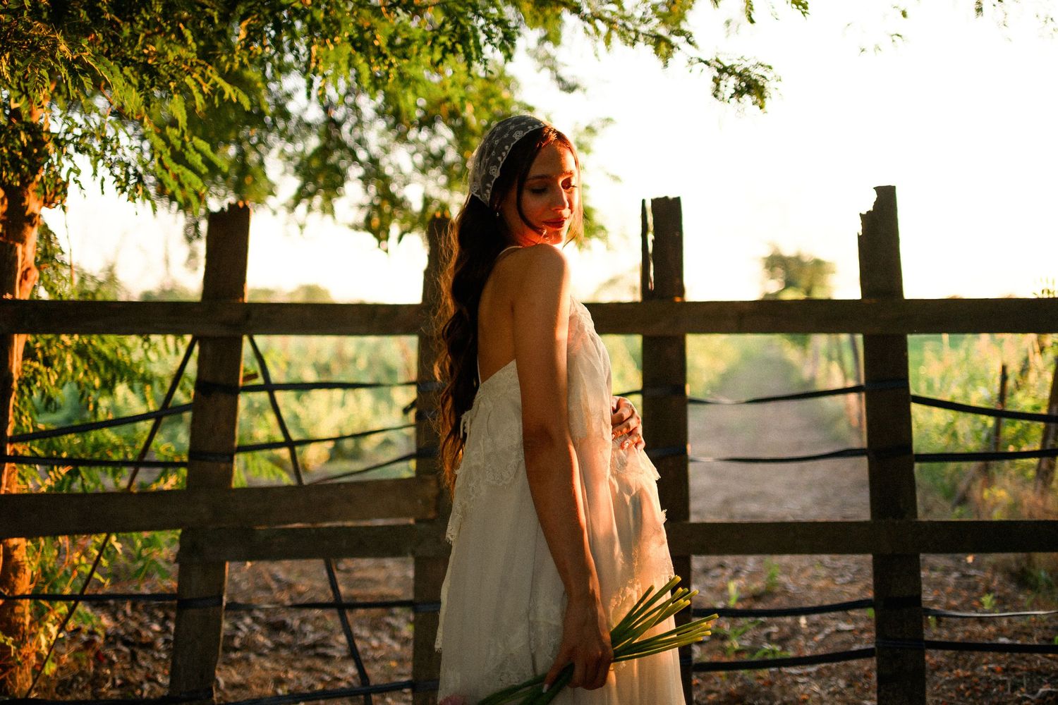 Woman in white dress standing by rustic wooden fence at golden hour sunset with lush green trees.