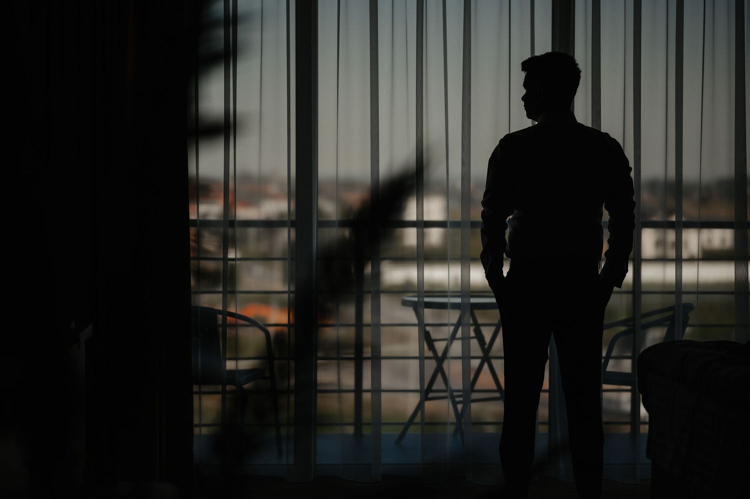 Silhouette of groom standing by large window with city view in dramatic dark lighting.
