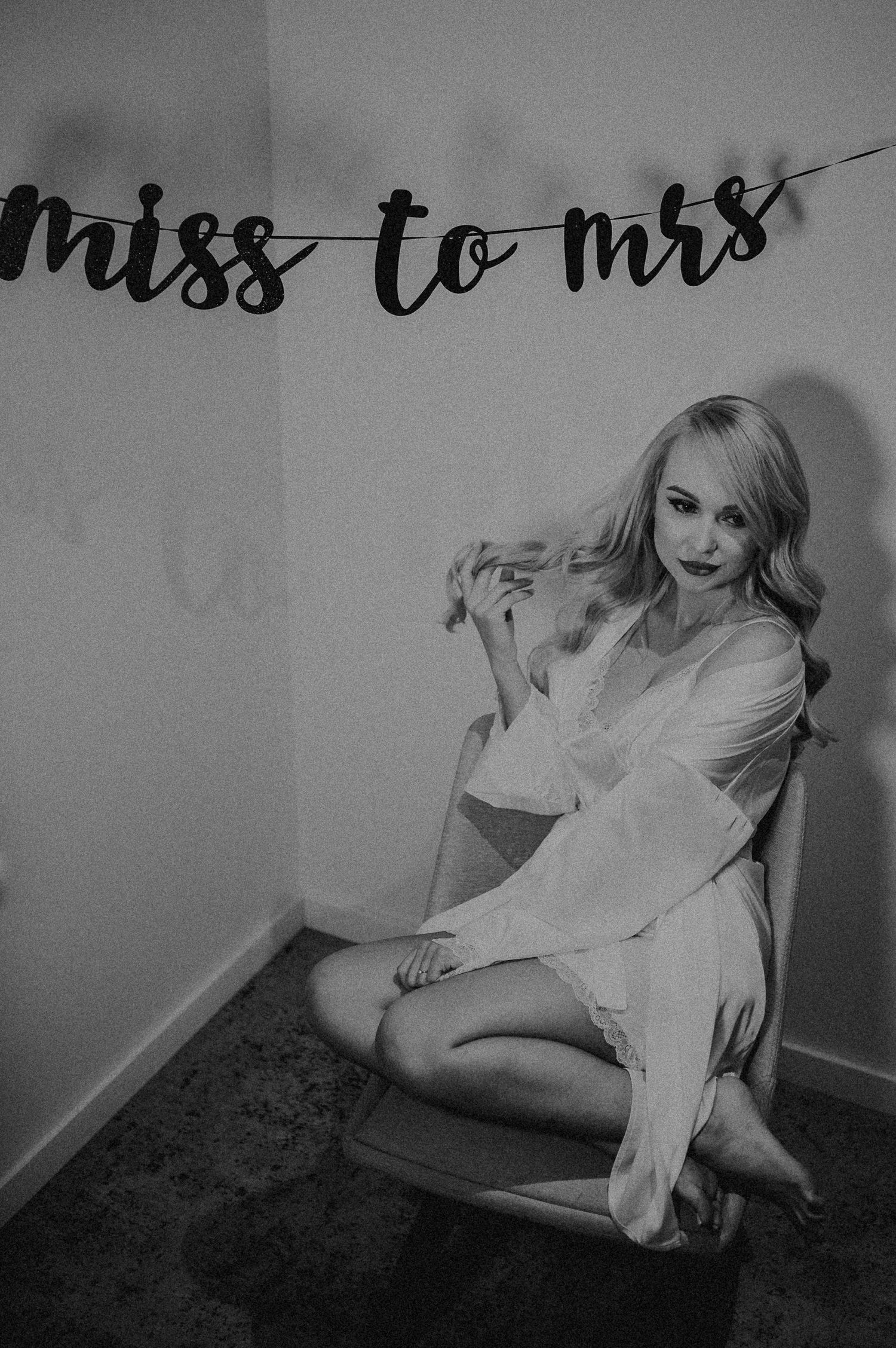 Blonde bride-to-be poses in white robe under miss to mrs banner in black and white boudoir photo.
