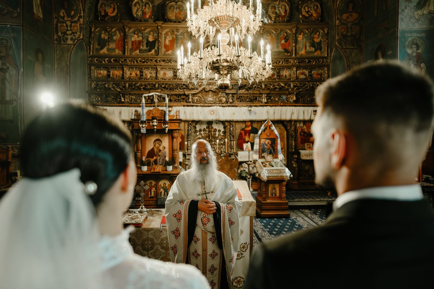 Bride and groom face Orthodox priest at ornate iconostasis during traditional church wedding ceremony.