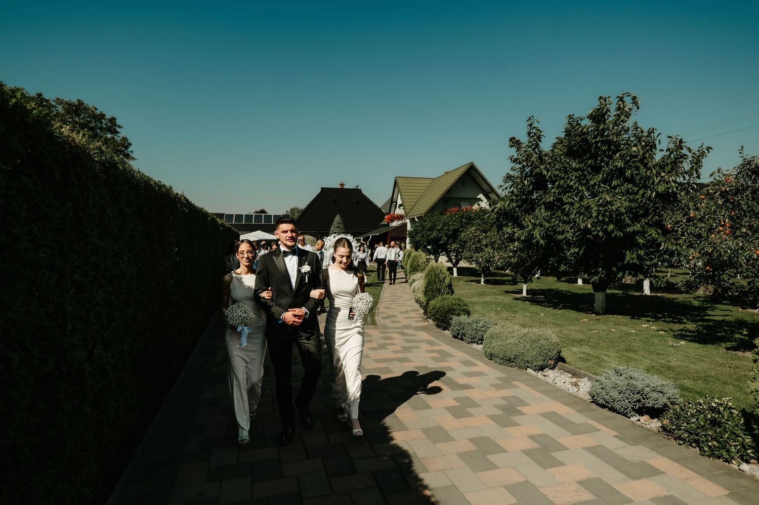 Wedding party walking along garden path on sunny day toward rustic venue.