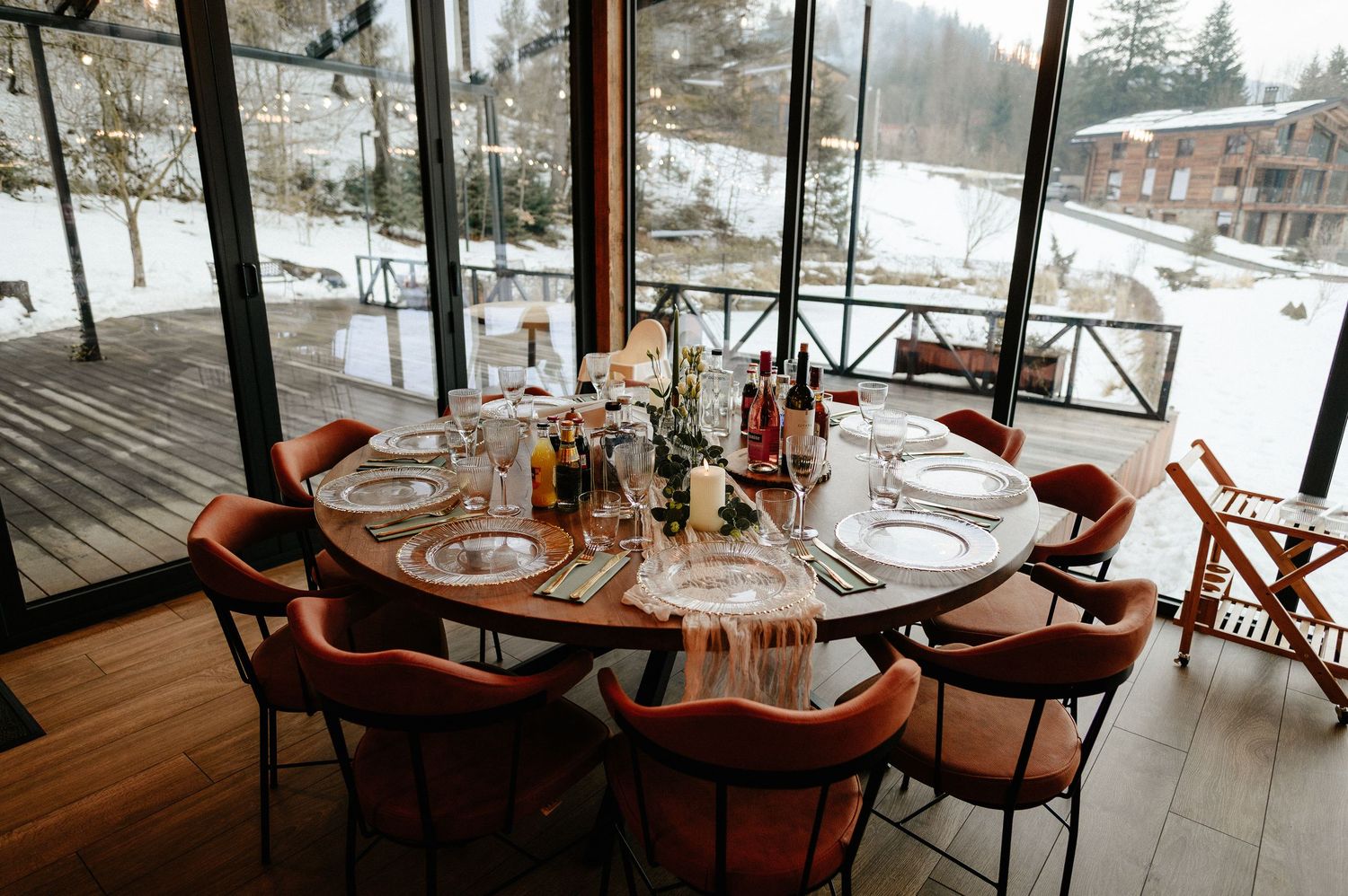Round dining table with rust-colored velvet chairs set for an intimate winter event with snowy views.