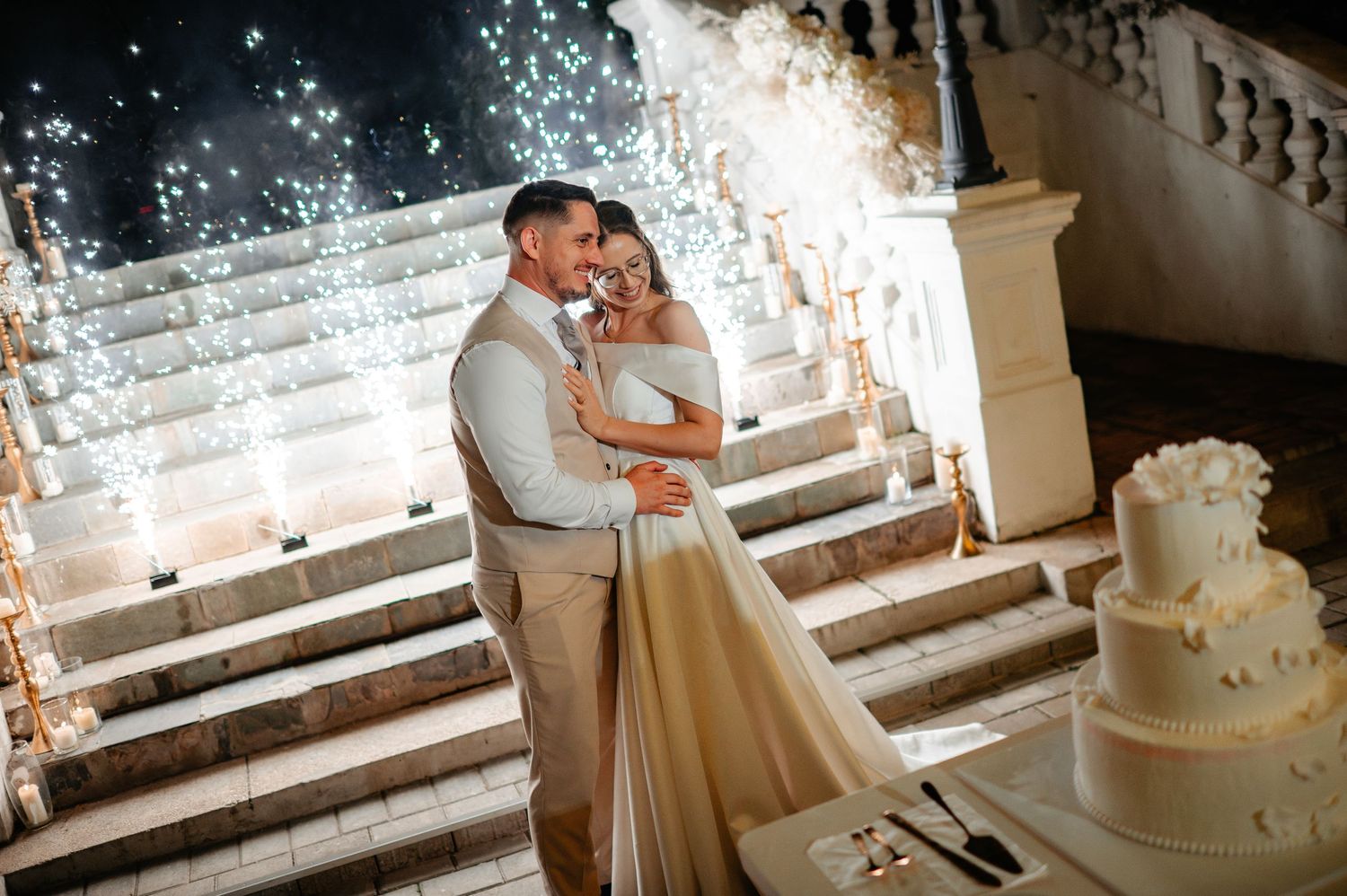 Newlyweds embracing near wedding cake with sparkling lights on grand staircase.