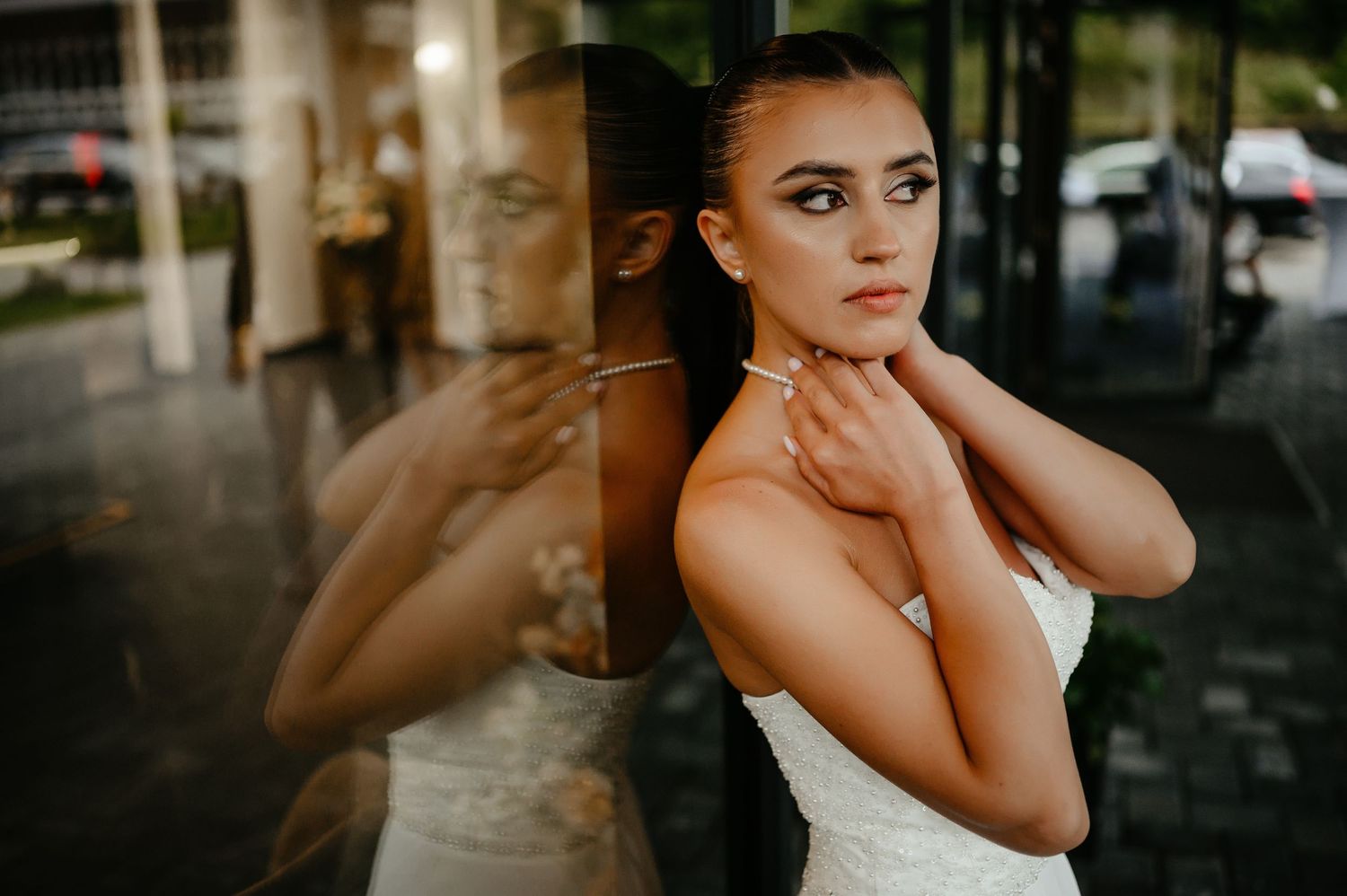 Elegant bride in a white gown poses against a glass window with a stunning reflection in natural light.