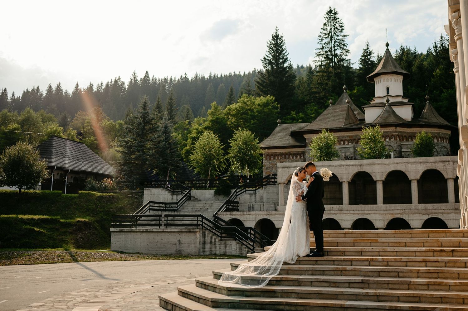 Wedding couple embracing on stone steps outside a stunning Orthodox monastery surrounded by lush pine forest.