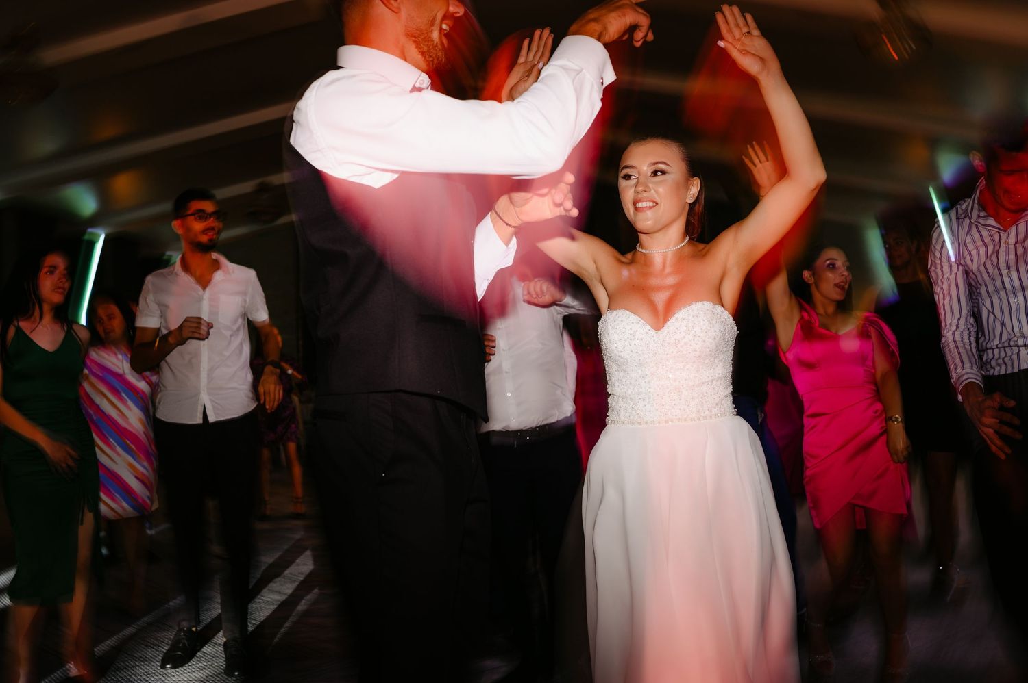 Smiling bride in white strapless gown dancing with groom at wedding reception with guests nearby.