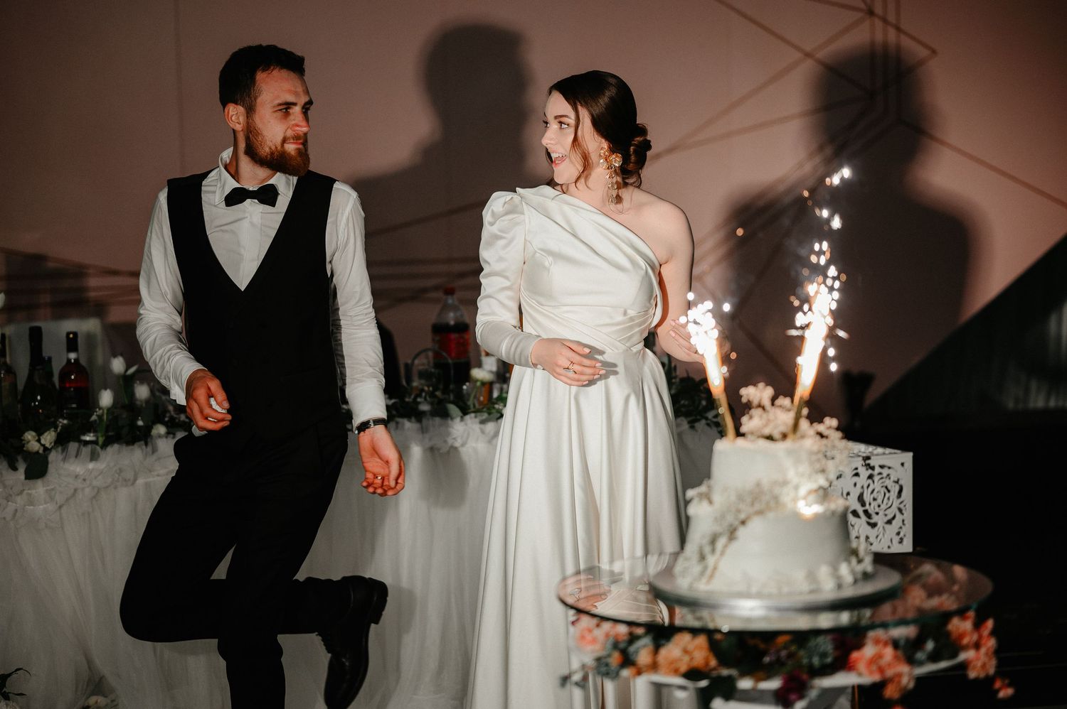 Bride and groom stand beside tiered white wedding cake with sparkling firework candles at reception.