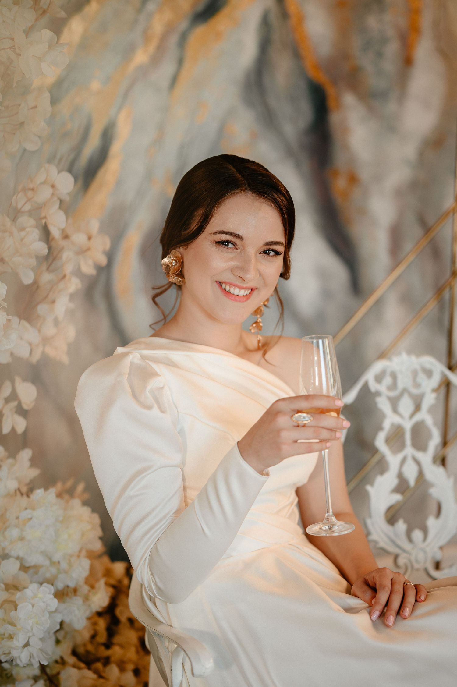 Smiling bride in white dress holds champagne glass while seated against decorative floral backdrop.