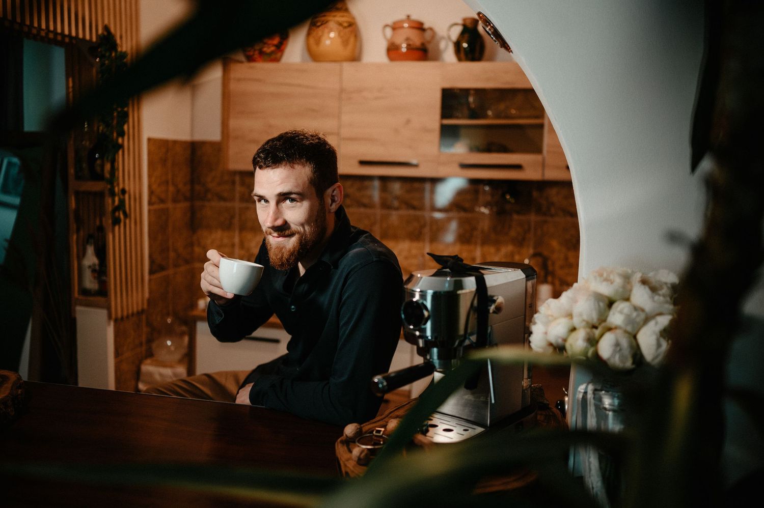 Bearded groom enjoying coffee at a wooden kitchen table beside an espresso machine and flowers.