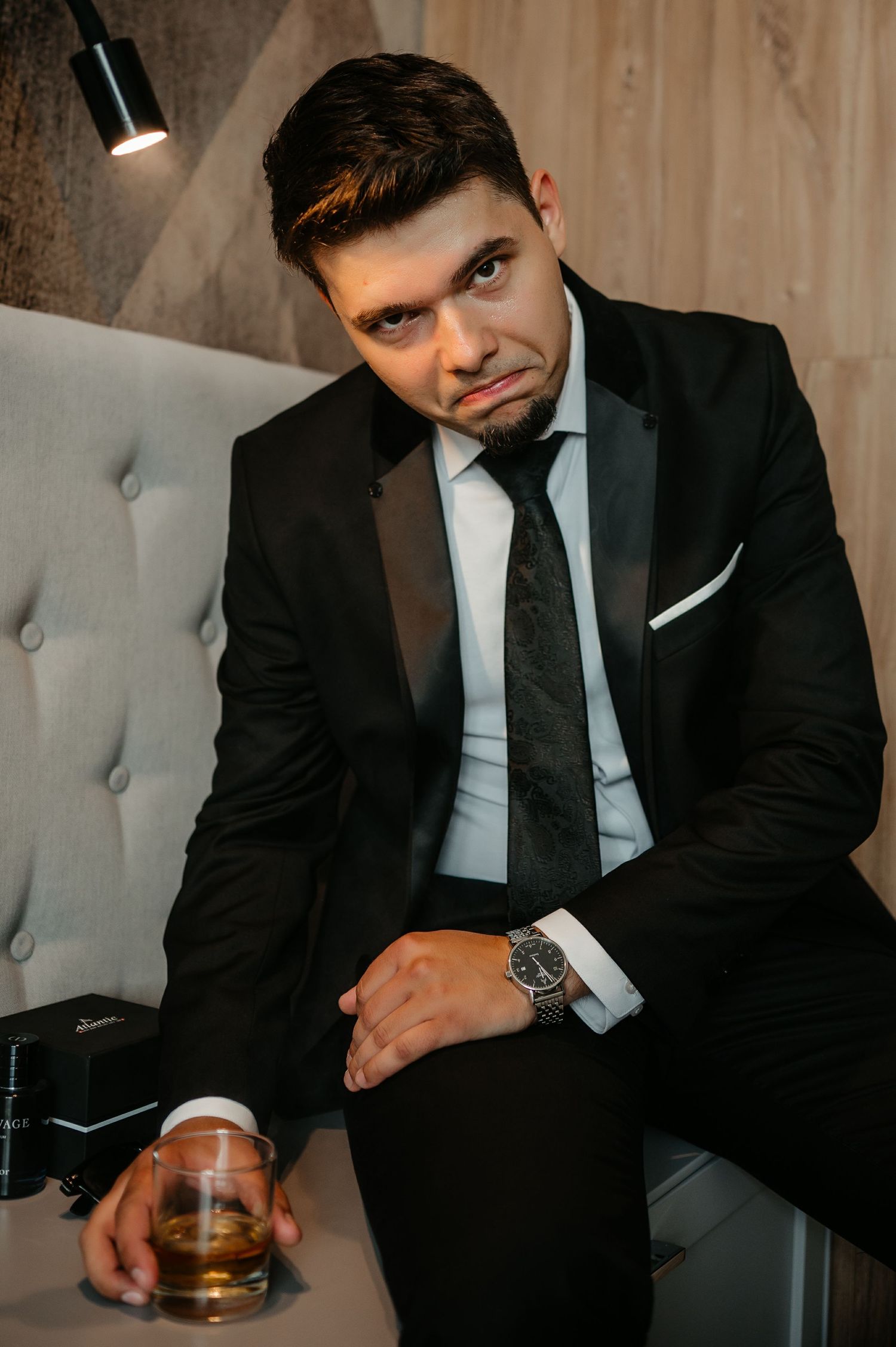 Stylish groom in black tuxedo sitting on couch holding whiskey glass with a confident expression.