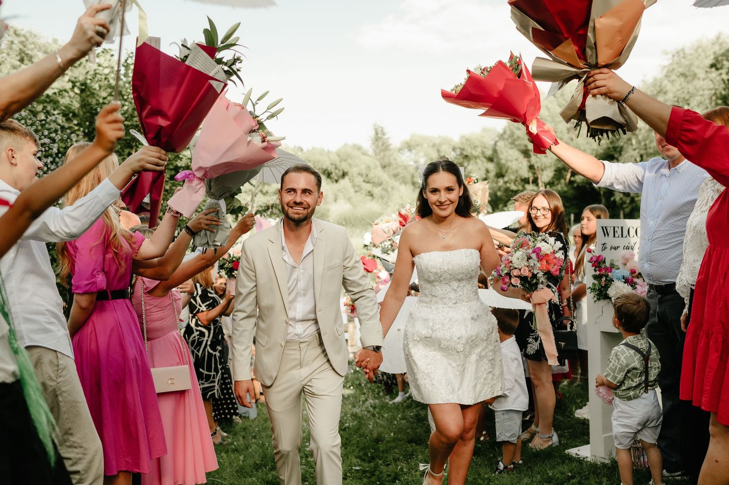 Newlyweds walk down the aisle as guests hold red flowers at an outdoor wedding ceremony.