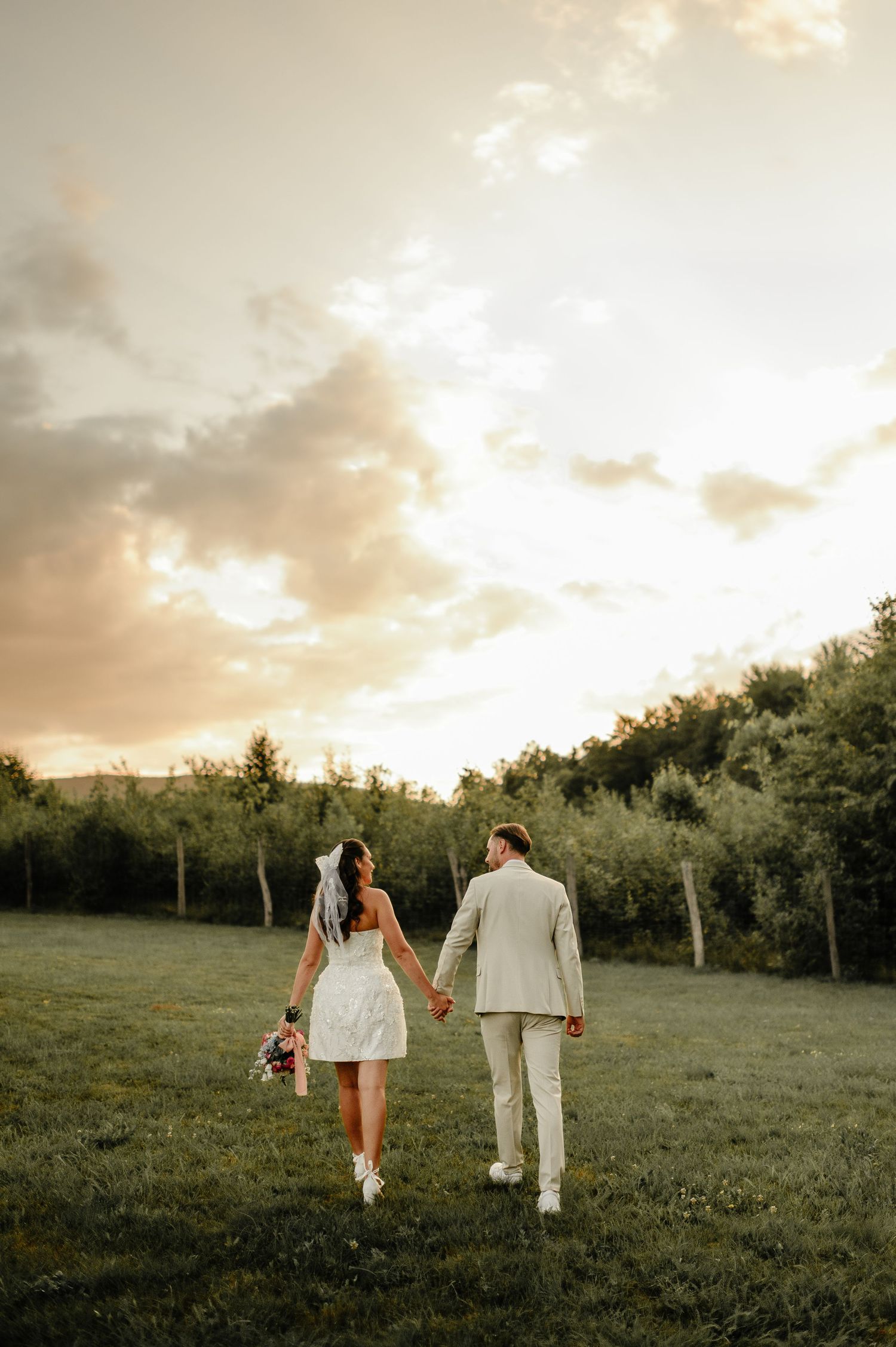 Bride and groom walking hand in hand across green meadow at golden hour sunset.