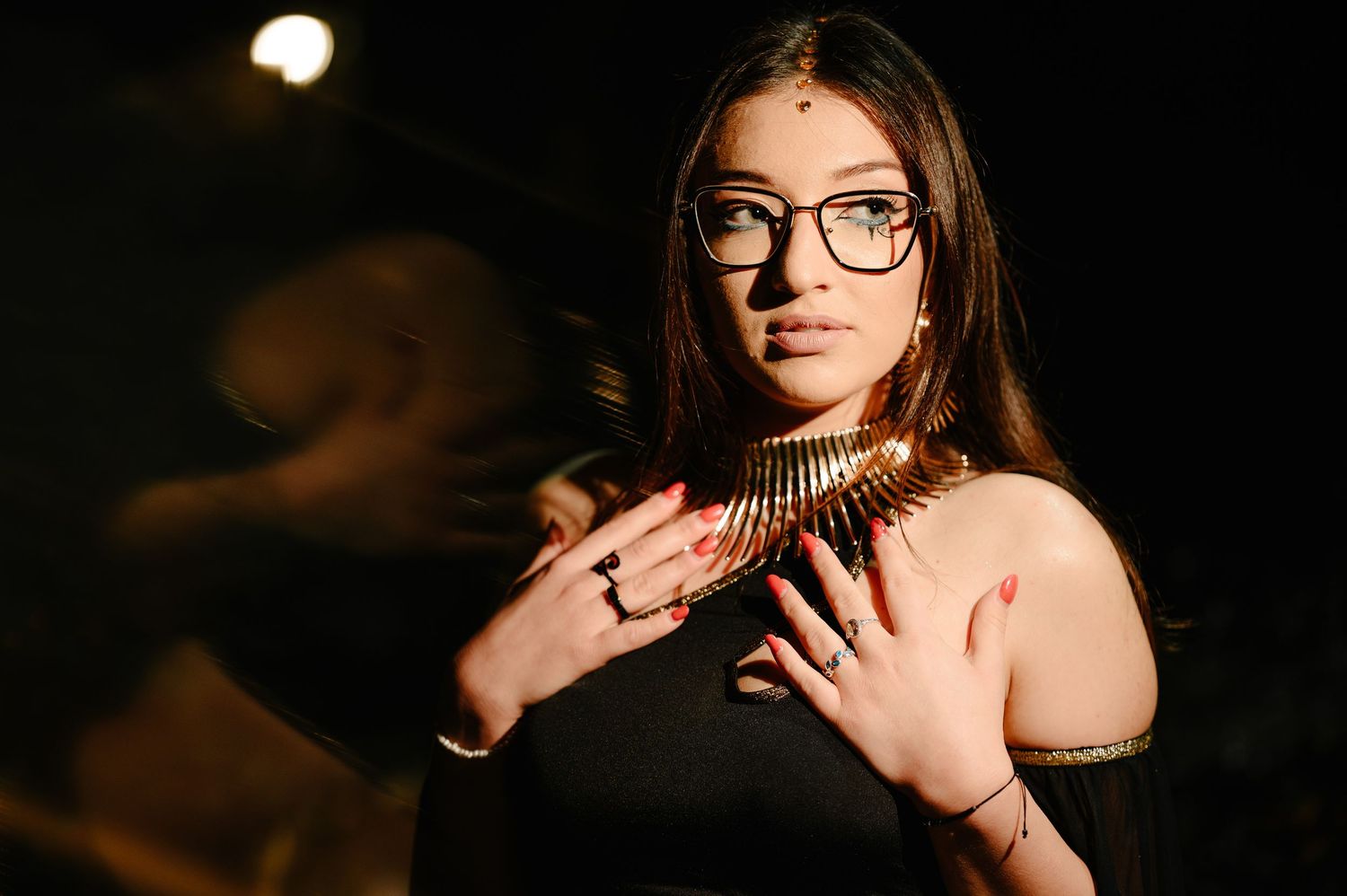 Young woman wearing glasses and gold collar necklace poses dramatically in dark setting.
