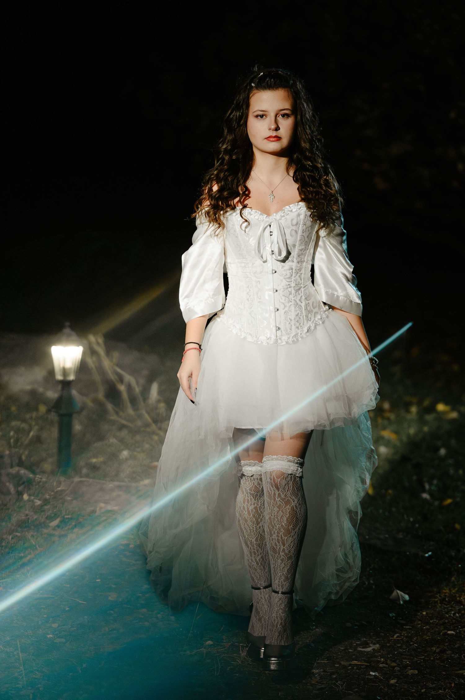 Young woman in white Halloween bride corset costume walking on dark path with street lamp and fog effect.