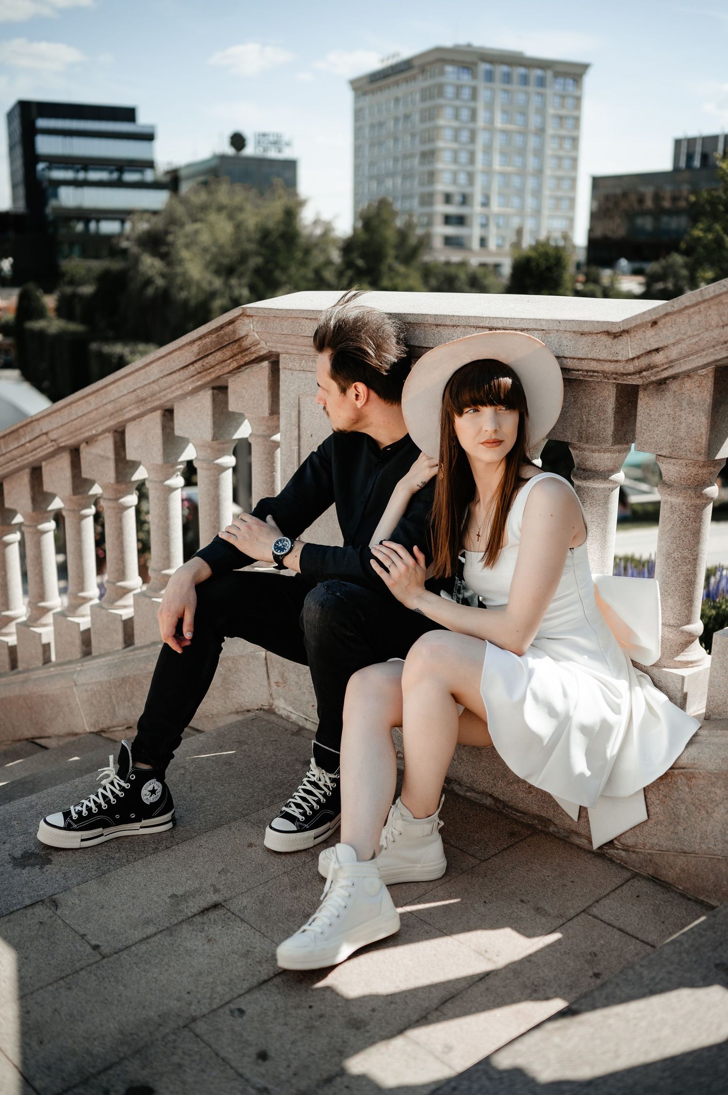 Stylish couple in contrasting black and white outfits sitting together on a stone balcony with city buildings behind.