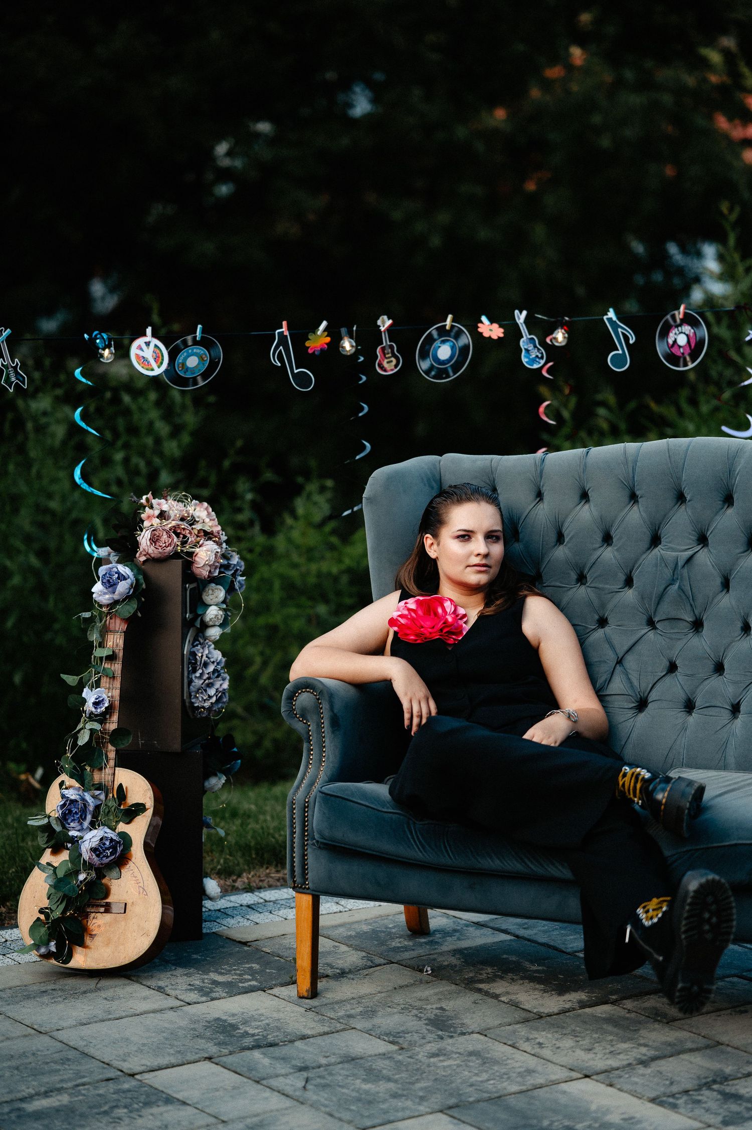 Young woman in black outfit with red flower sitting on gray sofa near a decorated guitar outdoors.