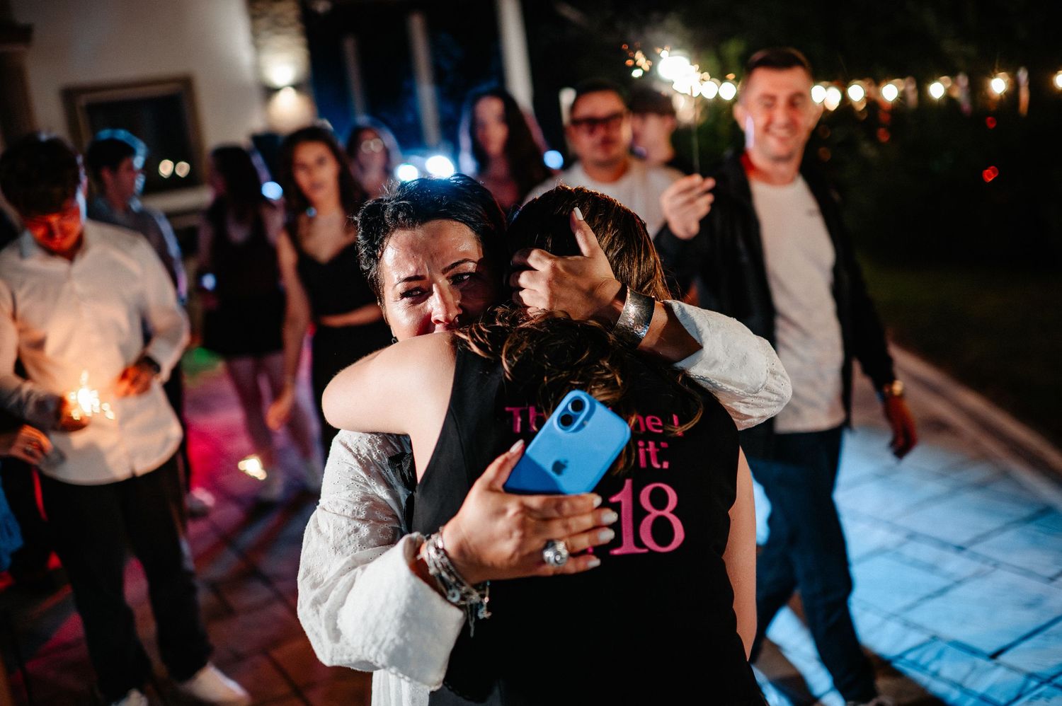 Two women share an emotional hug at an 18th birthday party with sparklers lighting the night.