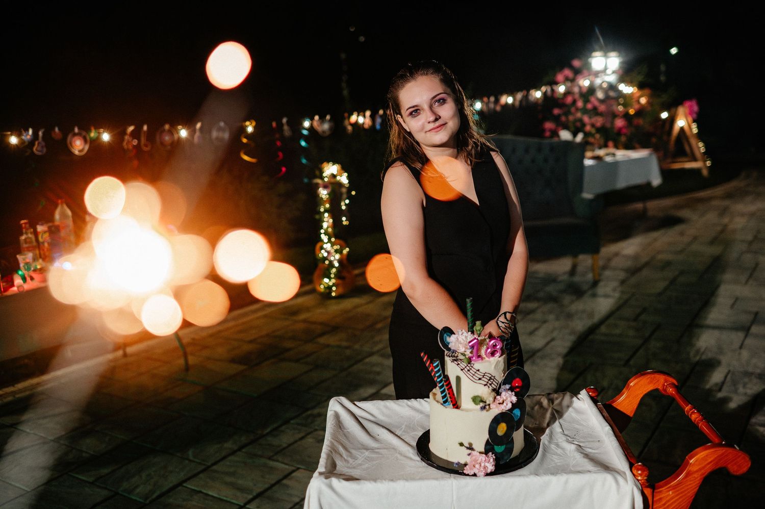 Young woman standing beside a decorated birthday cake at an outdoor night celebration with bokeh lights.