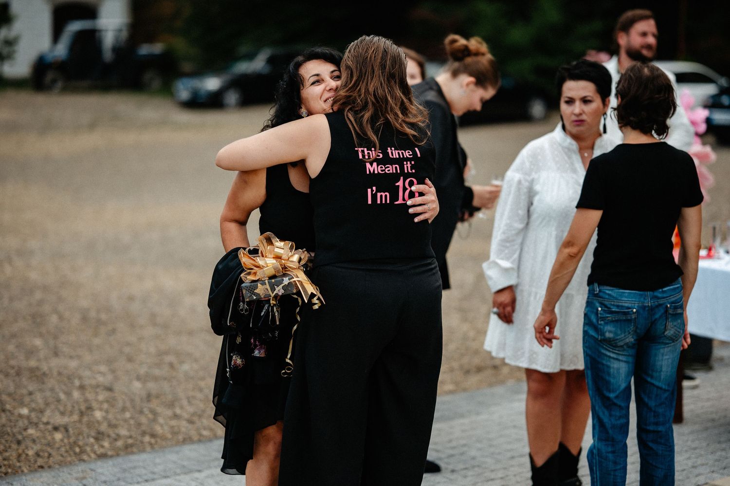 Two women embracing warmly at an outdoor event, one wearing a black jacket with text on the back.