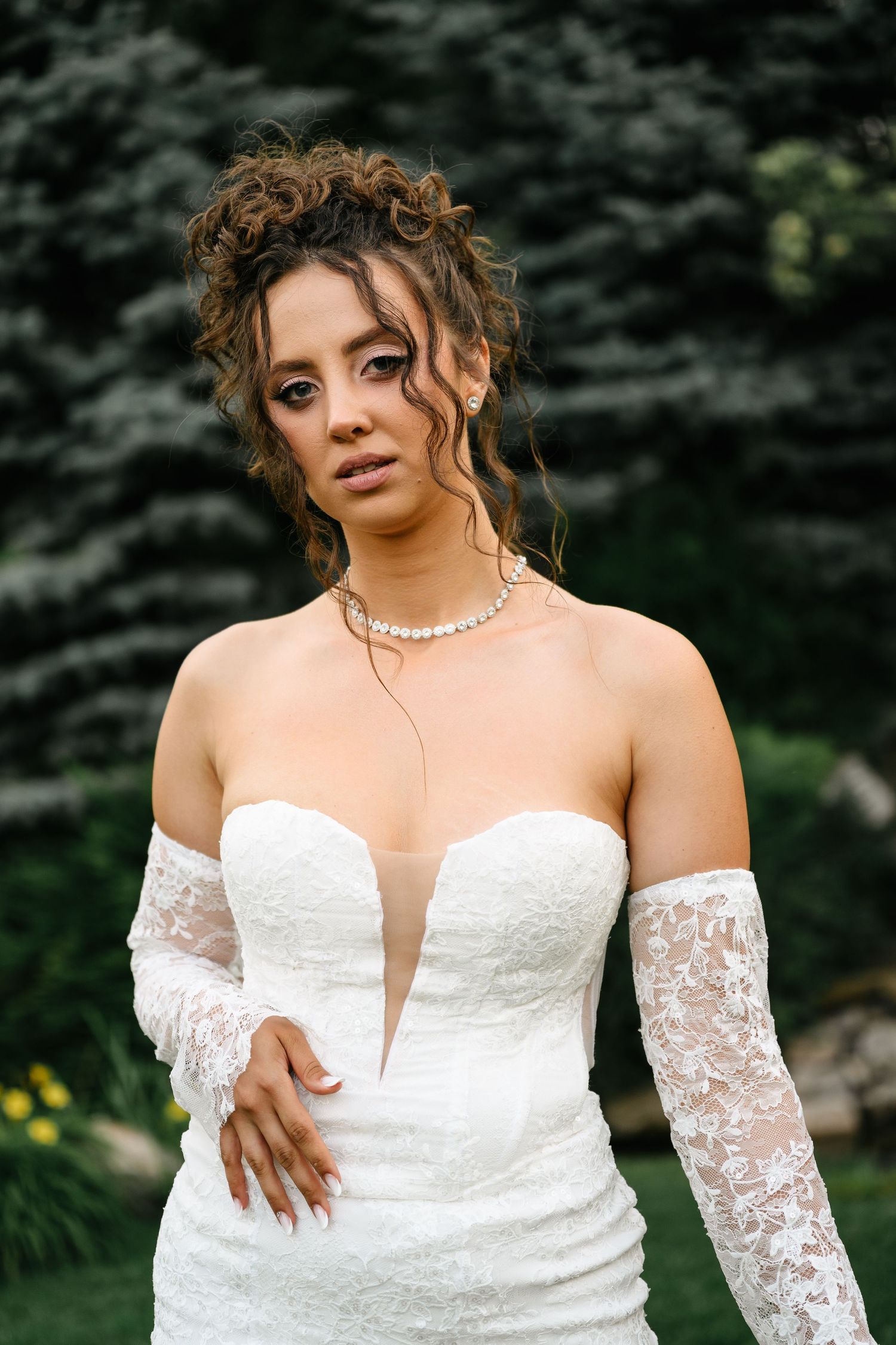 Bride in white lace off-shoulder gown with pearl necklace and updo hairstyle posing outdoors.