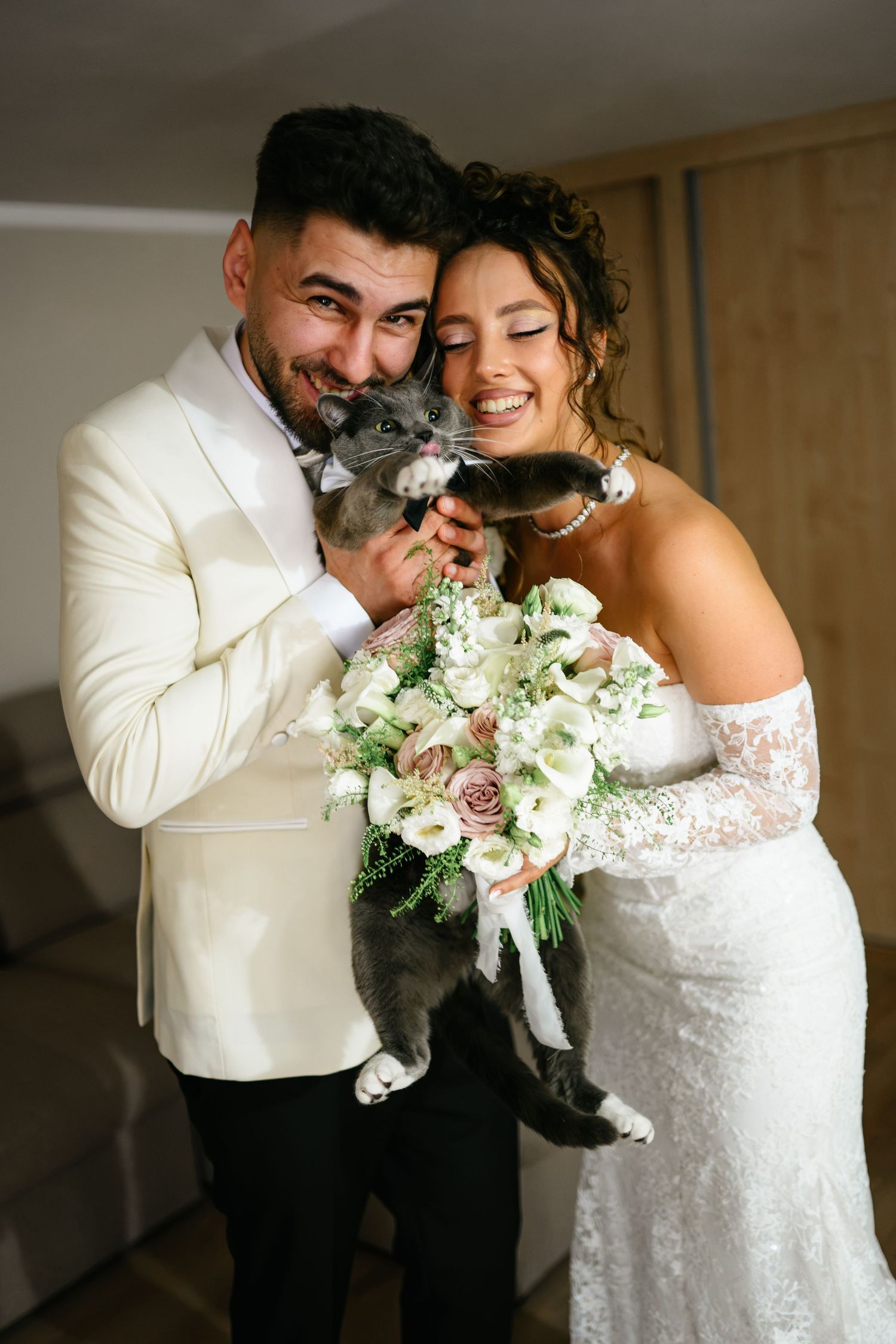 Smiling bride and groom in wedding attire holding a gray cat and bridal bouquet of white and pink flowers.
