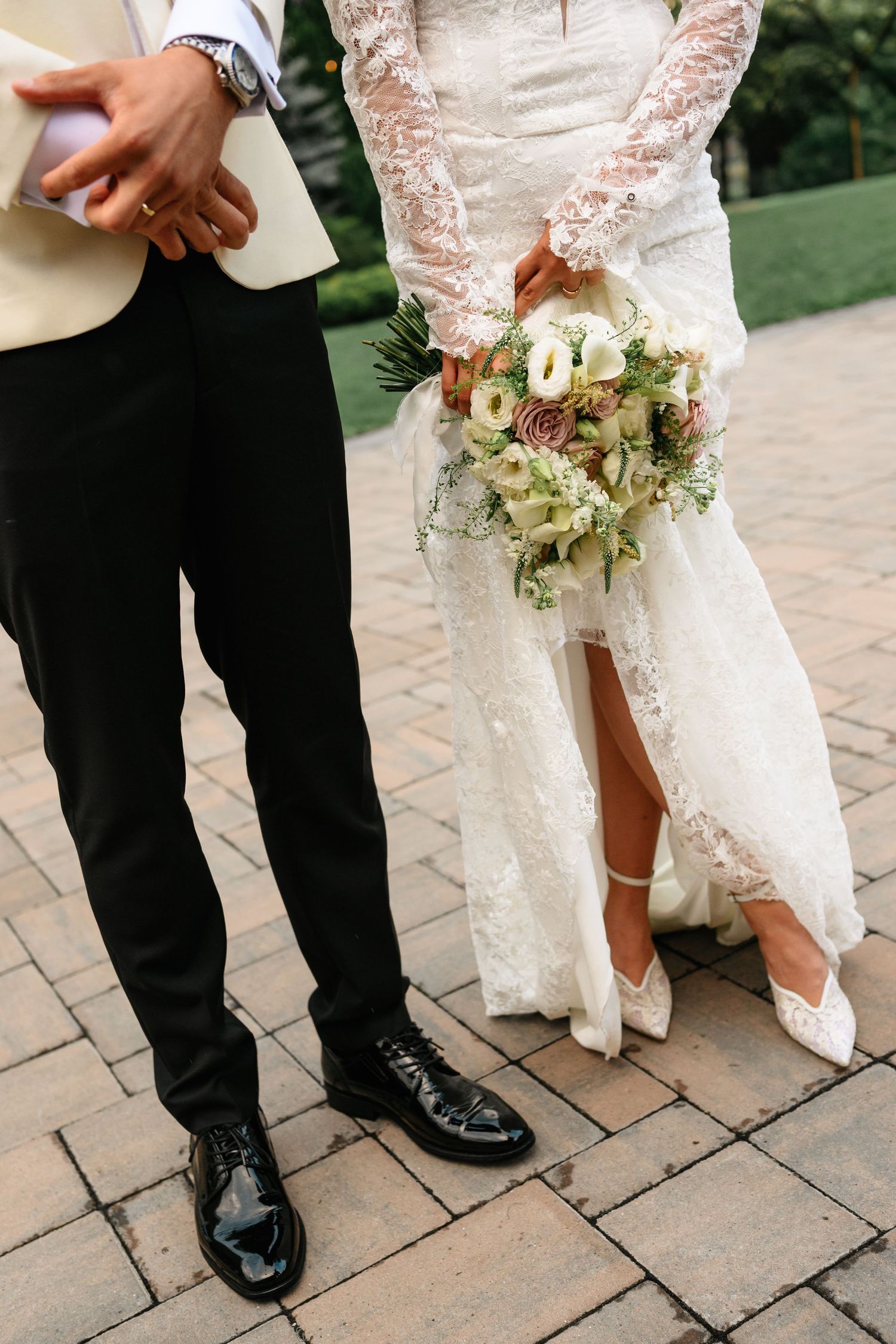 Bride in lace gown holding floral bouquet stands beside groom in black pants and shiny dress shoes on cobblestone.