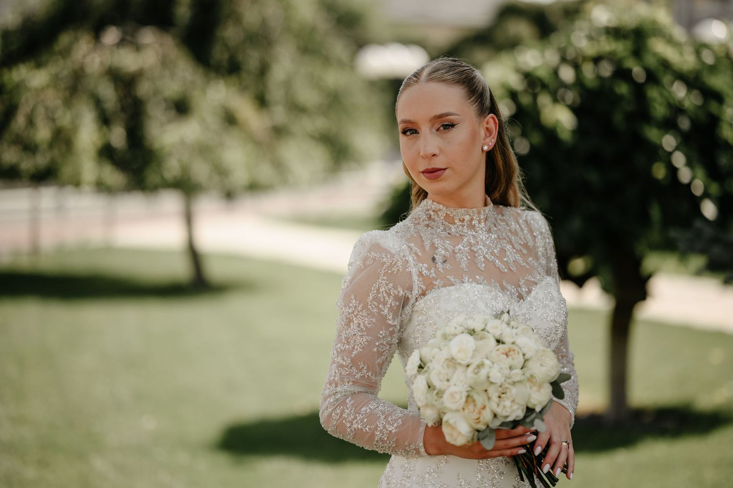 Beautiful bride in beaded high-neck long-sleeve wedding dress holding white bouquet outdoors.