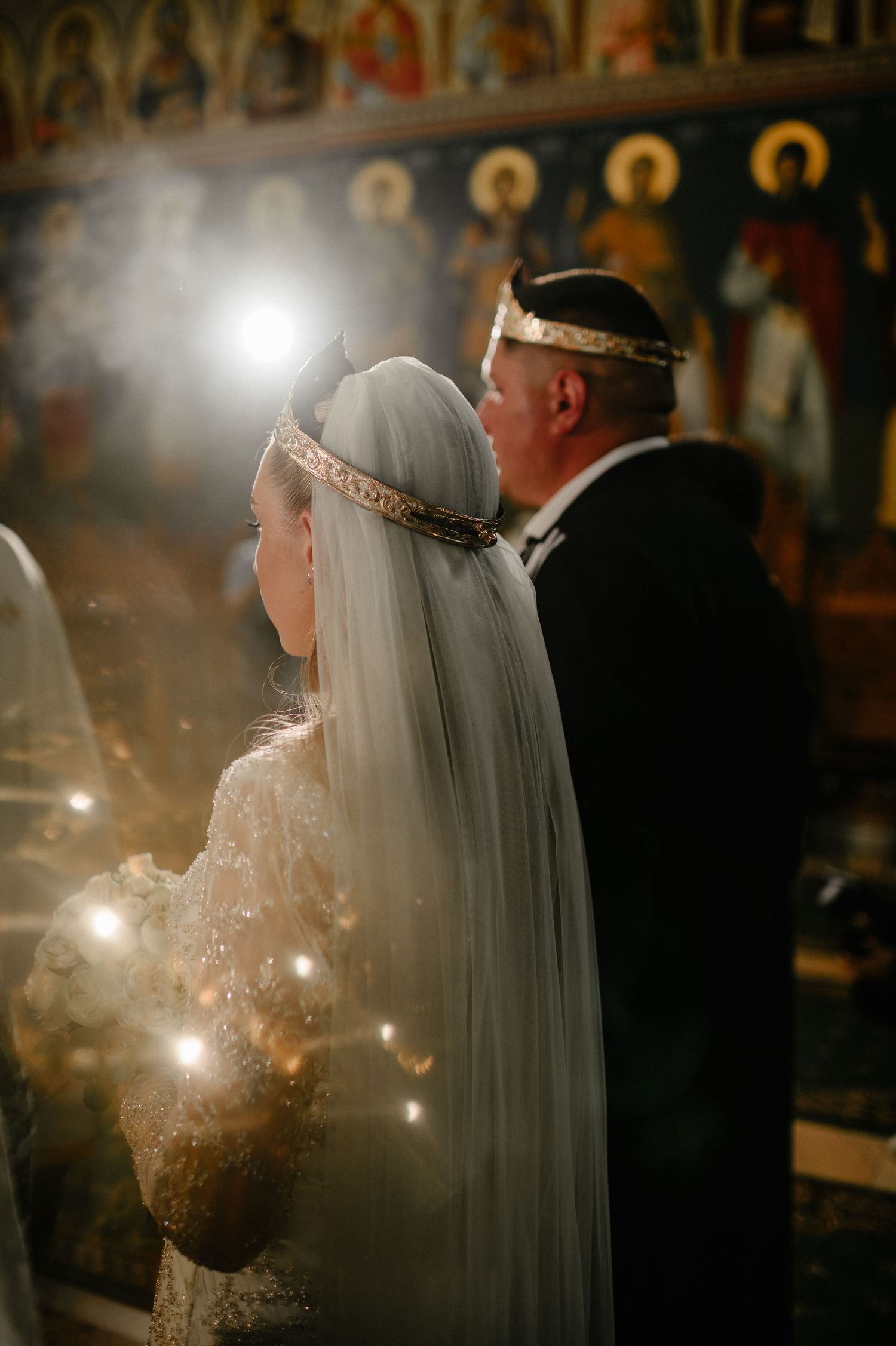 Bride and groom wearing orthodox wedding crowns stand before golden icons during a traditional Romanian church ceremony.