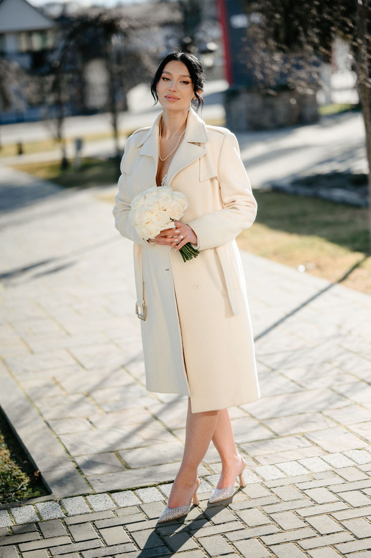 Elegant bride in cream coat holding white bouquet on sunny brick pathway.