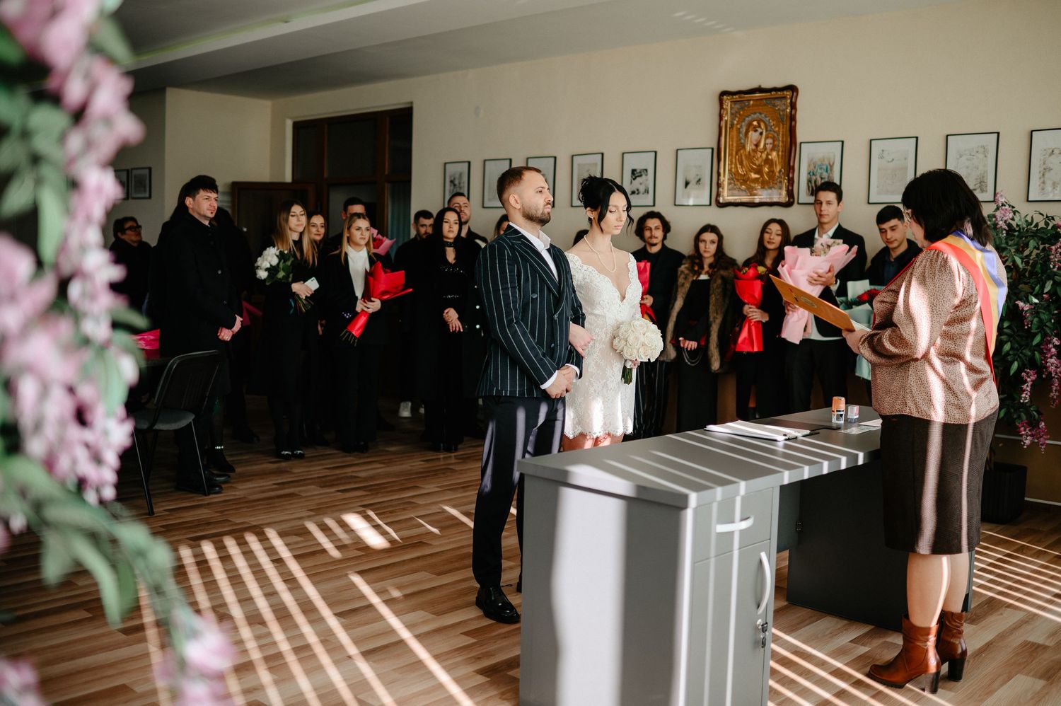 A couple exchanges wedding vows before an officiant in an elegant hall with guests seated in the background.
