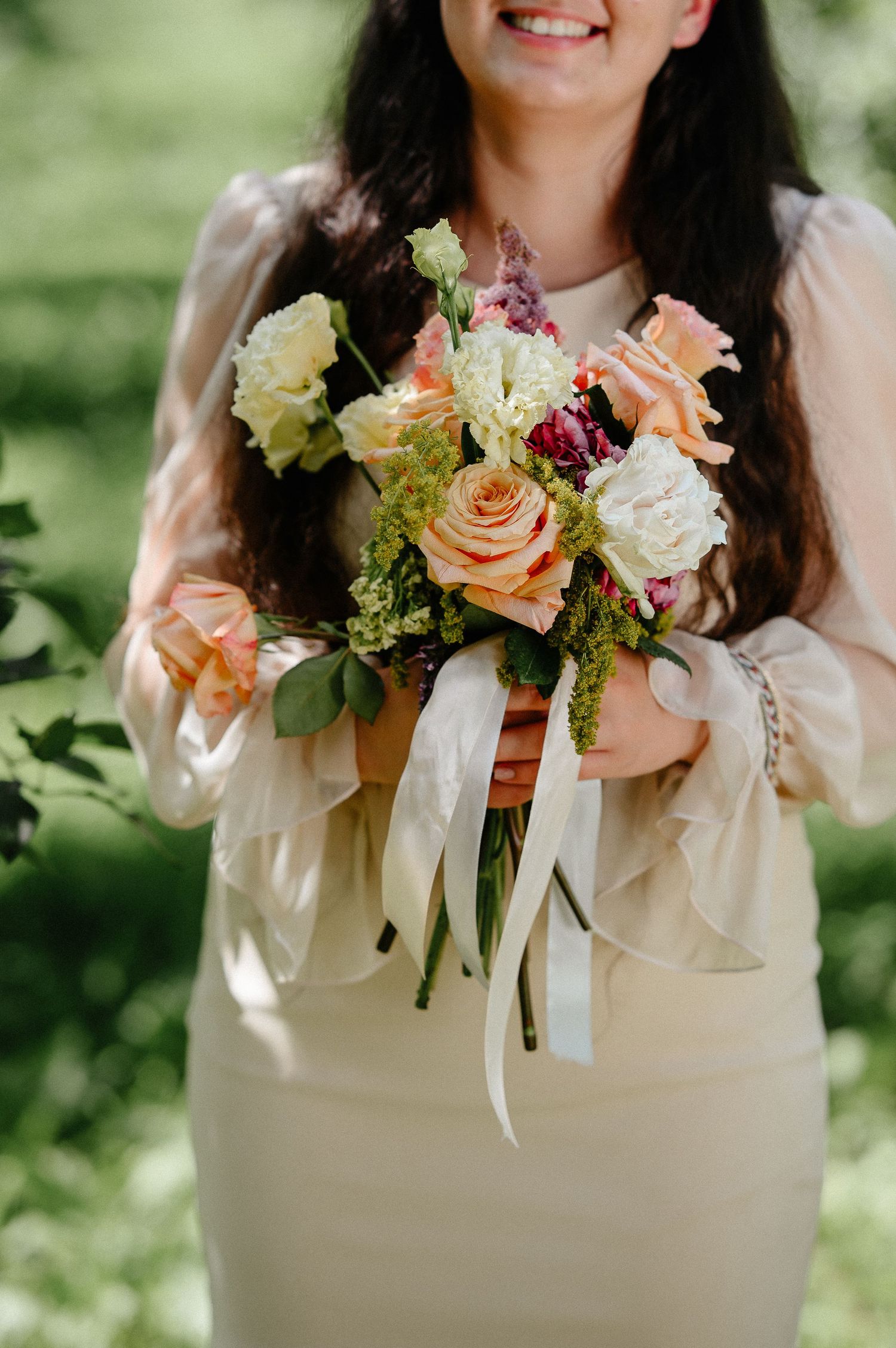 Smiling bride holding a lush bouquet of peach roses and white flowers with ribbon streamers outdoors.