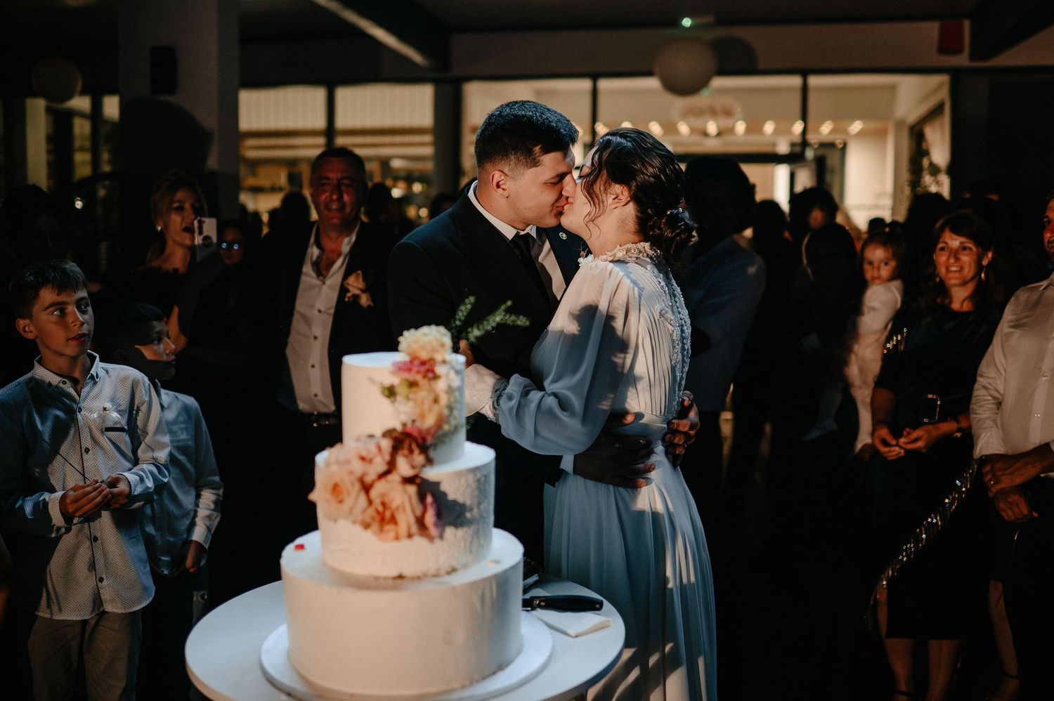 Bride and groom kiss beside three-tier wedding cake at lively evening reception.