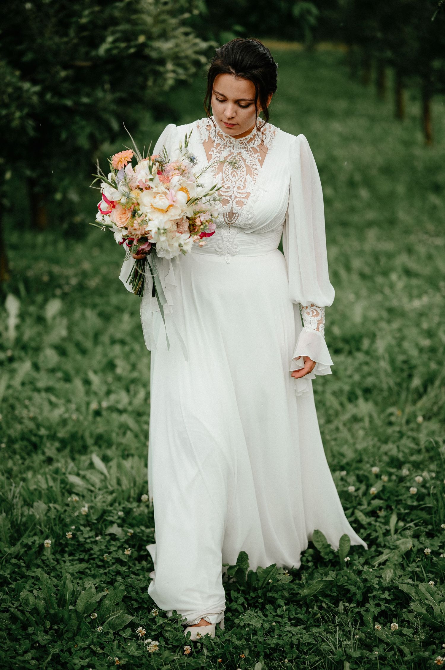 Bride in elegant white long-sleeve lace wedding gown holding colorful bouquet outdoors.