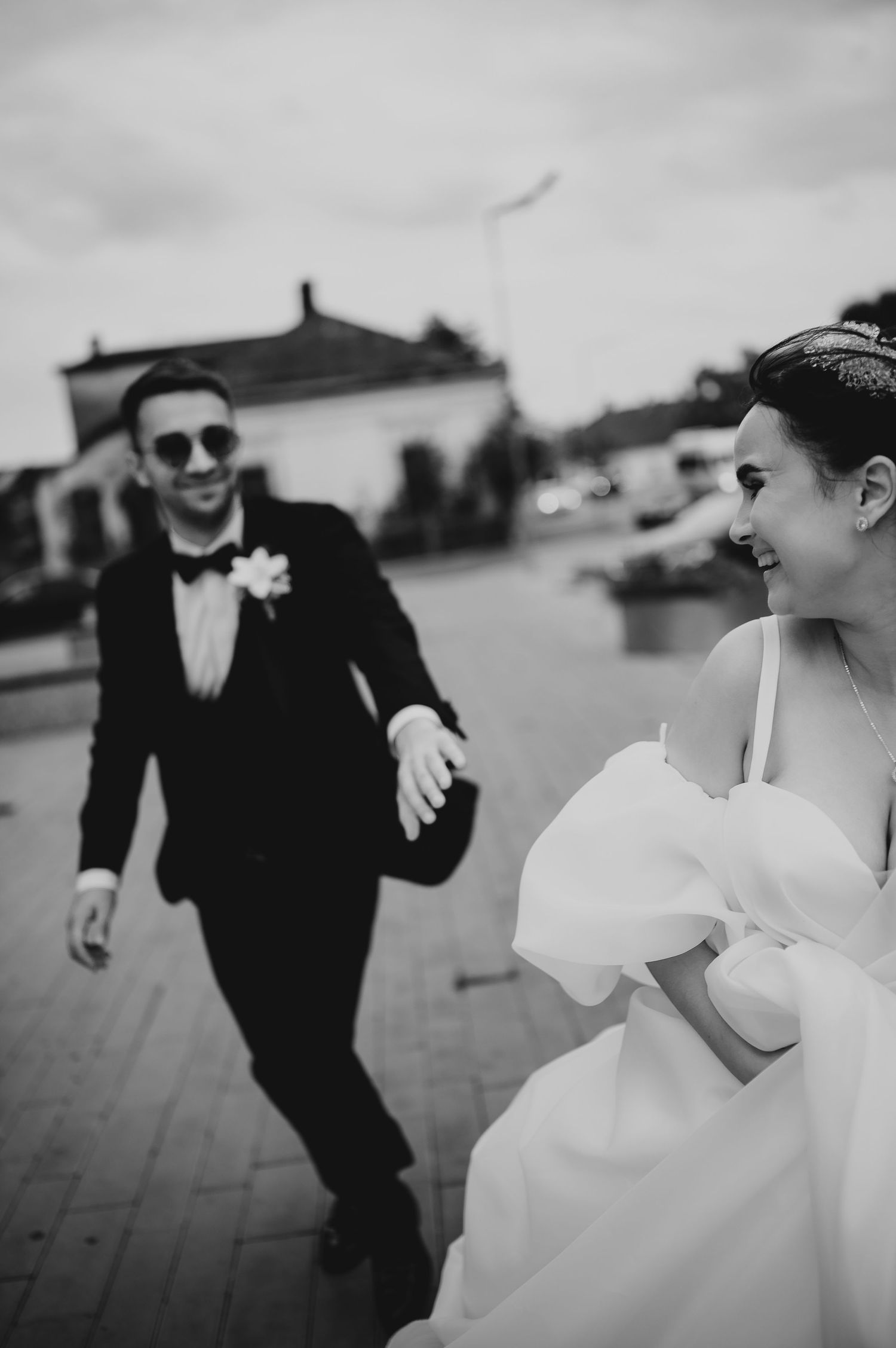 Black and white photo of smiling bride and groom in tuxedo holding hands running outdoors on wedding day.
