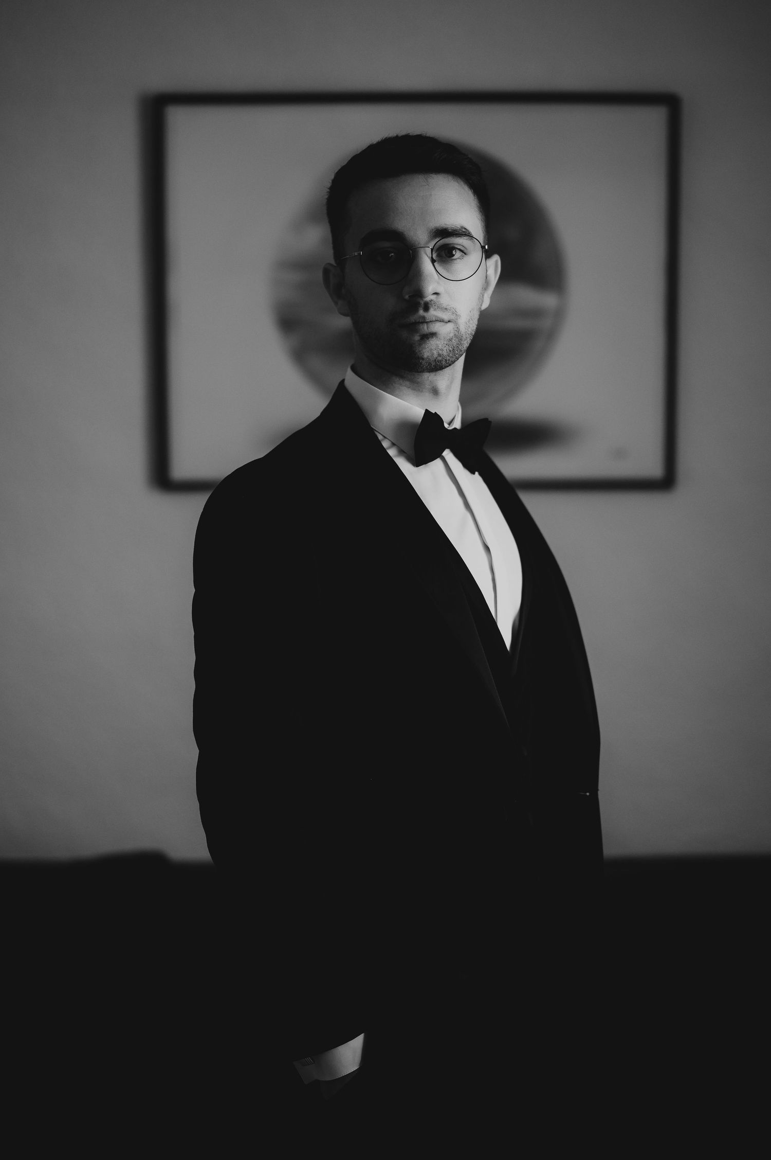 Elegant man in black tuxedo and bow tie wearing glasses poses in black and white portrait.