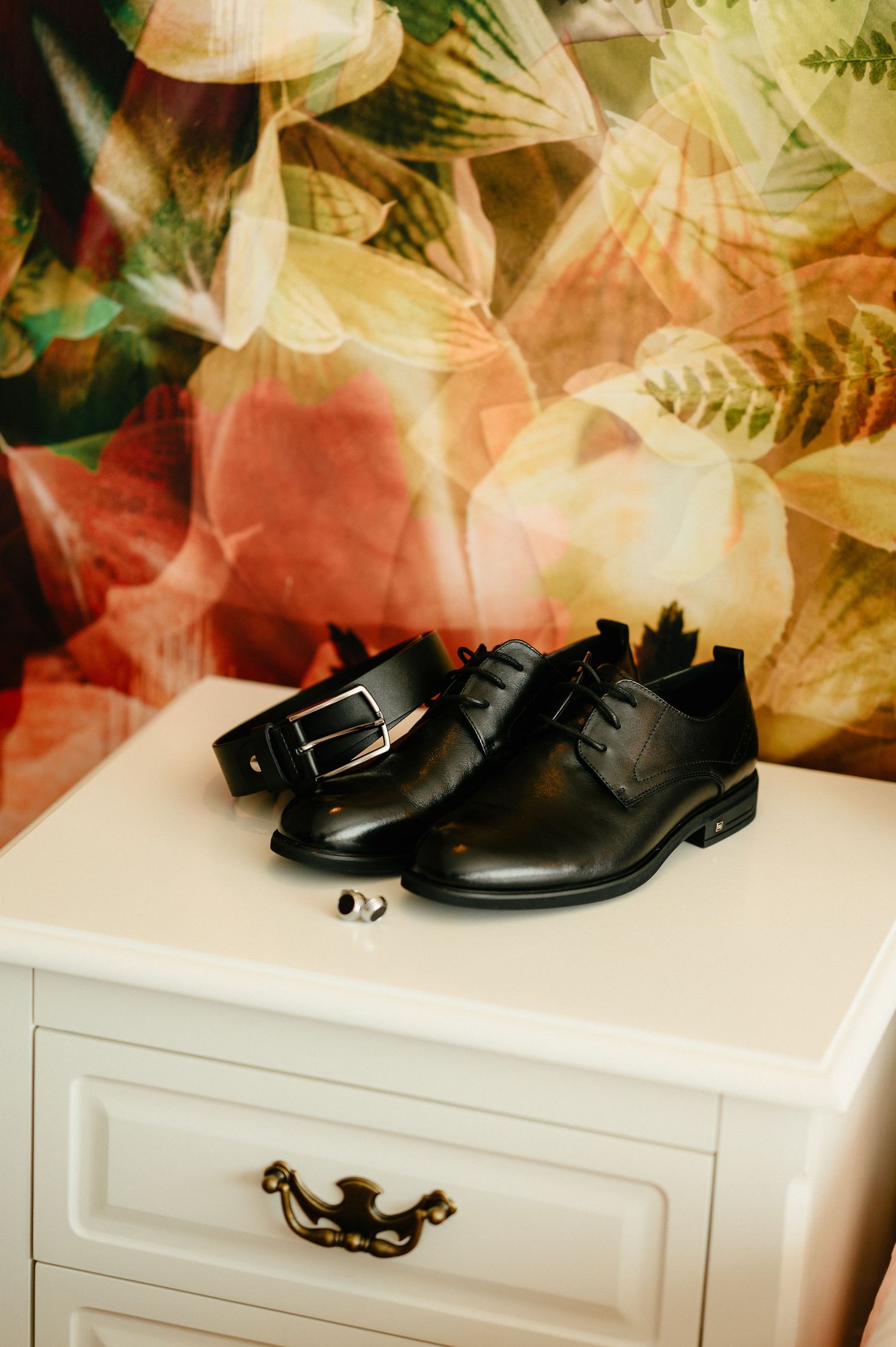Black leather dress shoes and belt on white dresser with floral wallpaper background, perfect for formal occasions.