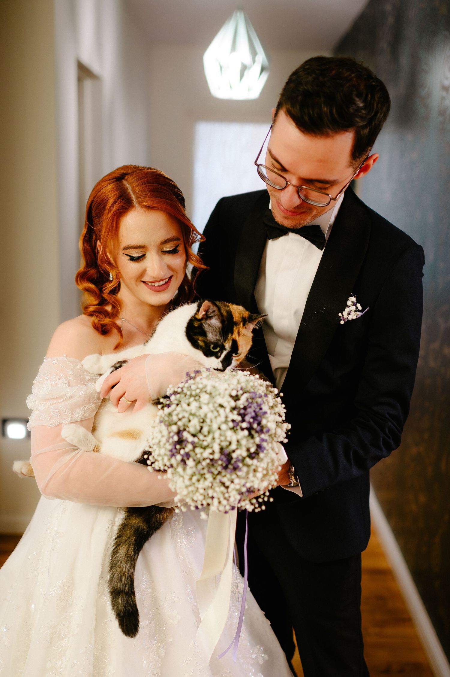 Bride and groom in wedding attire holding a calico cat and a bouquet of baby's breath and lavender flowers.