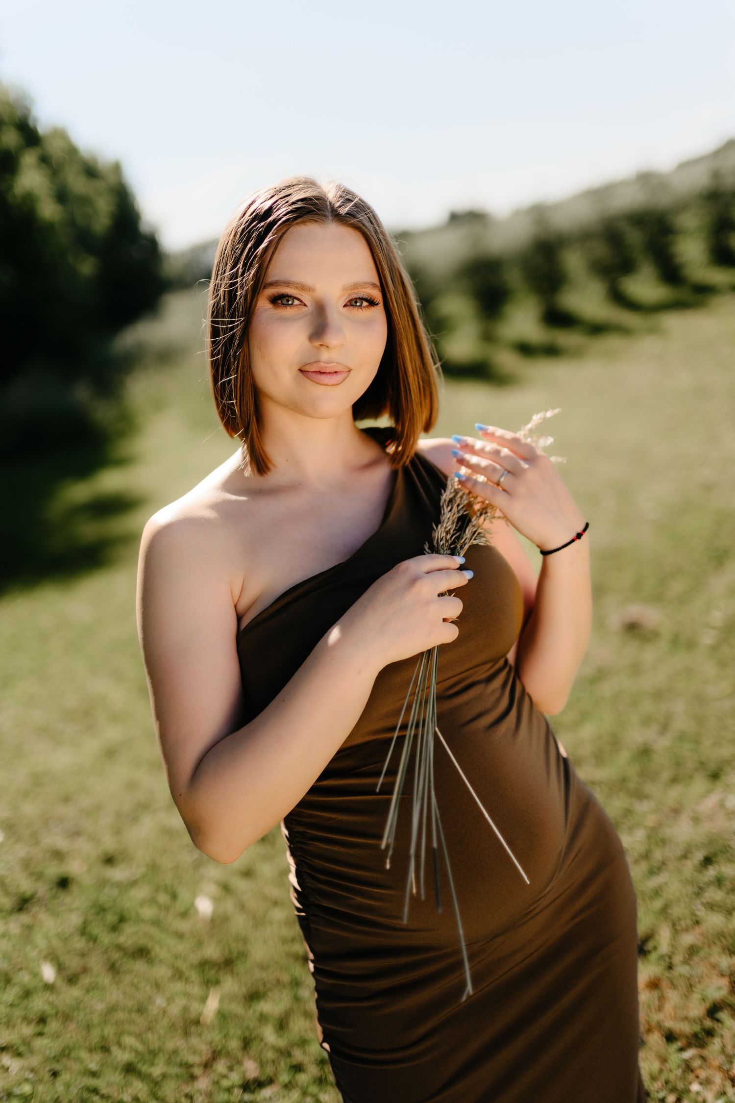Young woman in olive one-shoulder dress holding wildflowers in sunny green field.