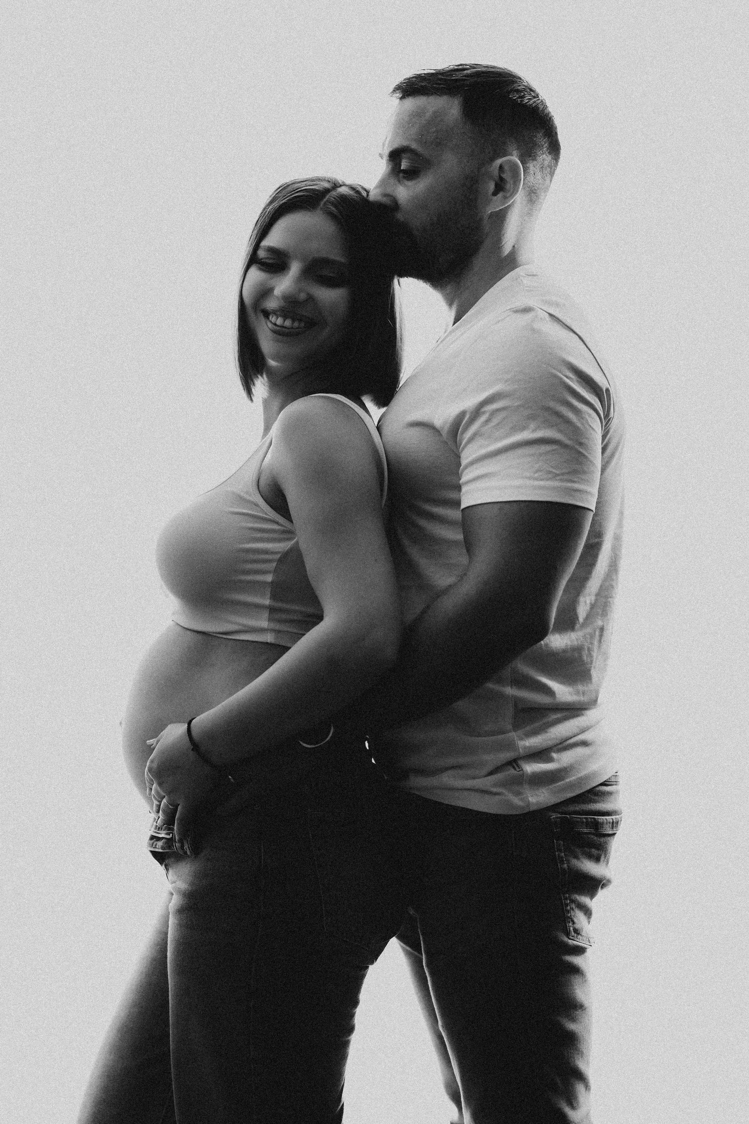 Black and white photo of a smiling pregnant couple embracing tenderly, man kissing woman's head from behind.