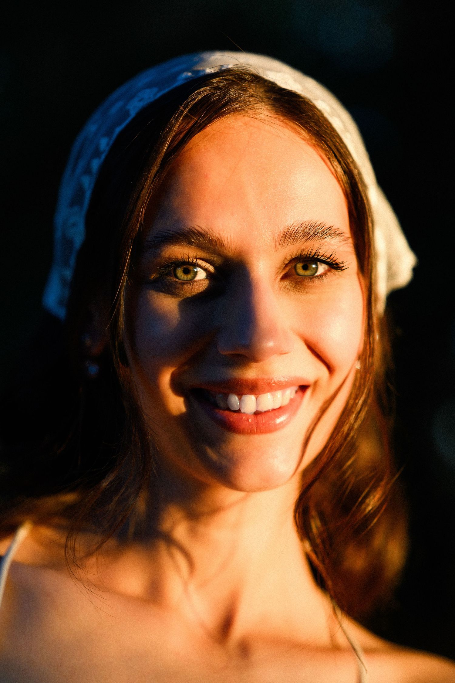 A smiling young woman with green eyes wearing a white headband, bathed in warm golden light against a dark background.
