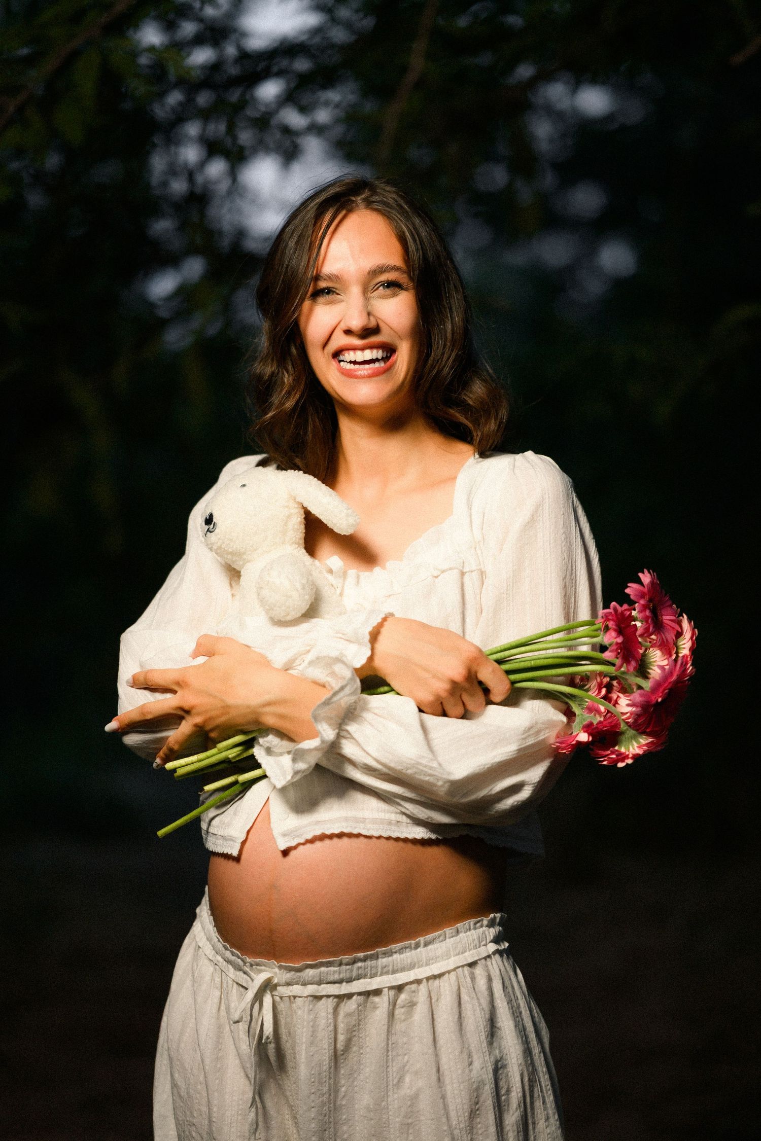 Smiling pregnant woman holding a white stuffed bunny and red flowers outdoors in natural light.