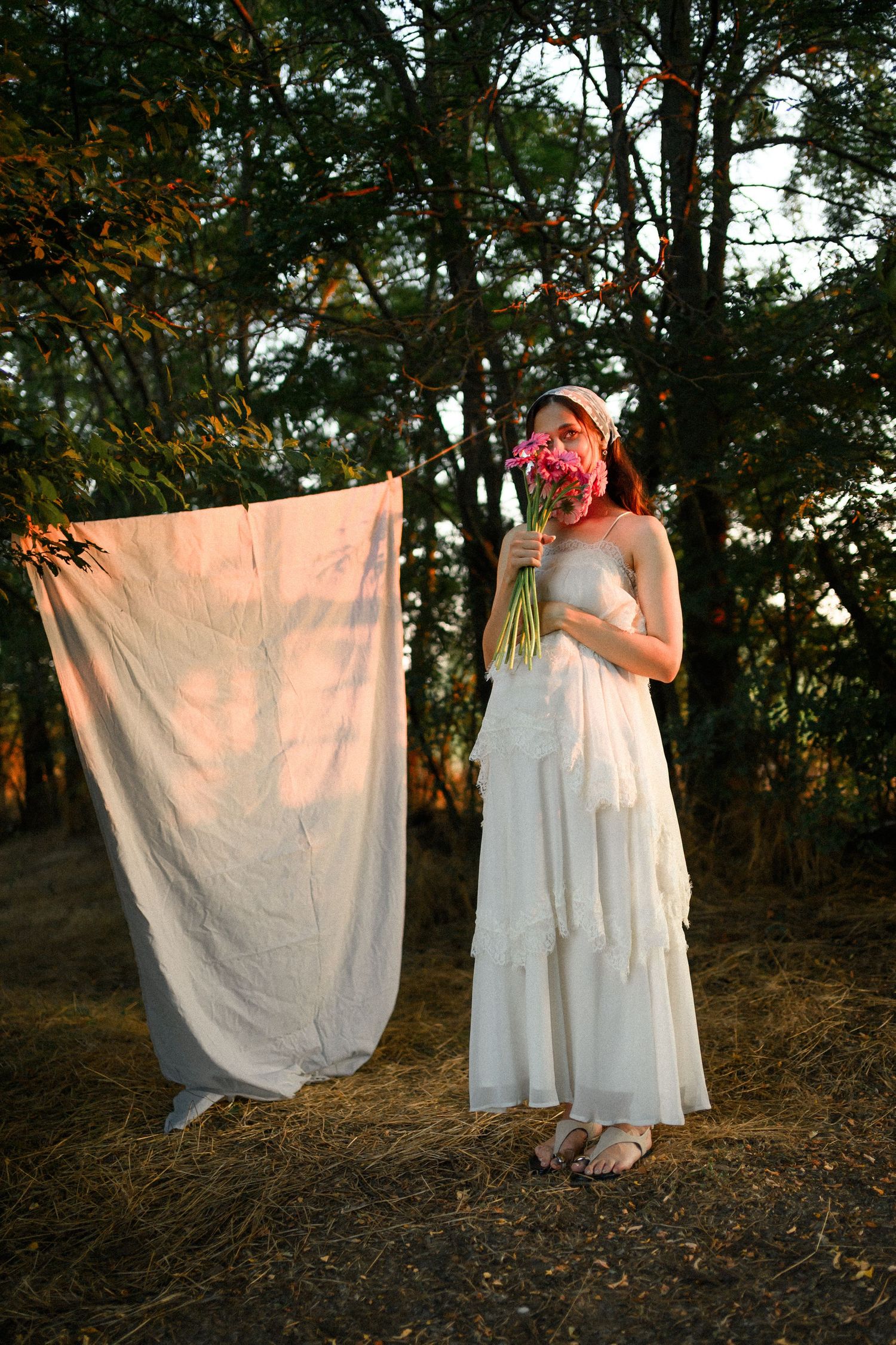 Woman in white dress and hat holds pink flowers near hanging linen cloth in a sunlit forest setting.