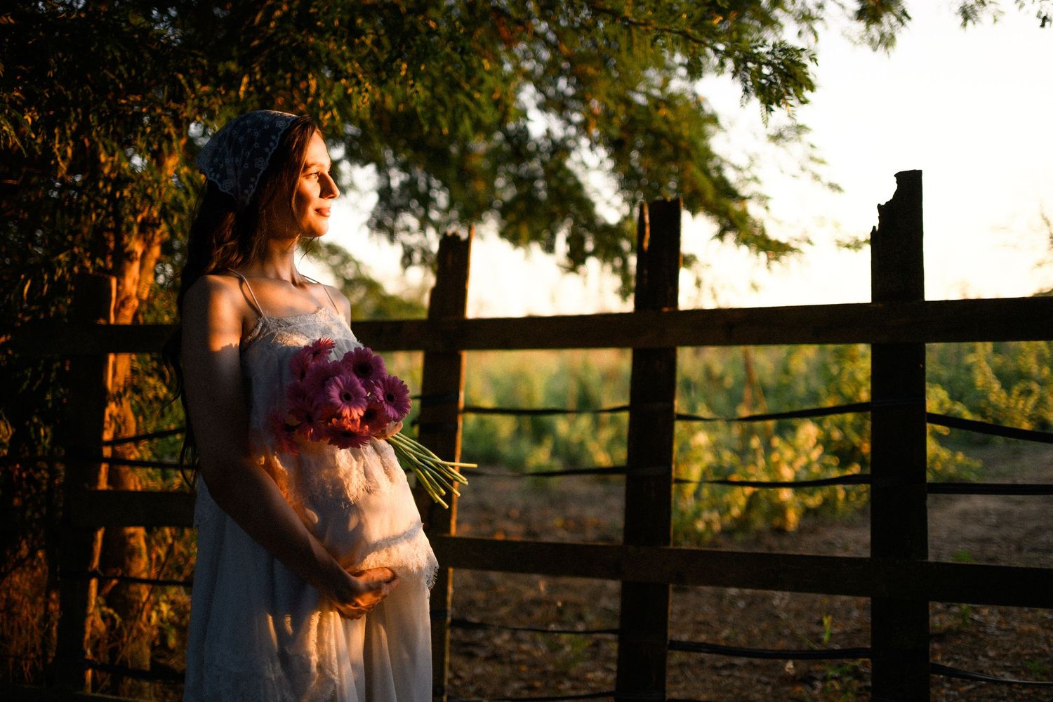 Pregnant woman in white dress holding purple flowers by wooden fence at golden hour sunset.