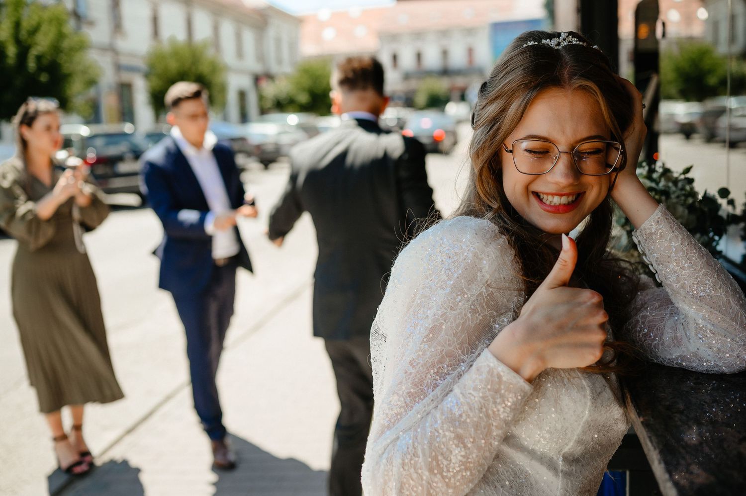 Happy bride in white lace dress and glasses giving thumbs up while guests celebrate behind her.