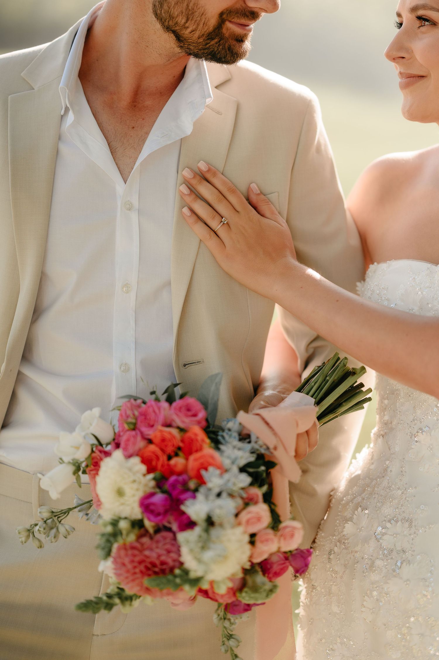 Bride holding colorful bouquet places hand on groom's chest in beige suit during outdoor wedding ceremony.