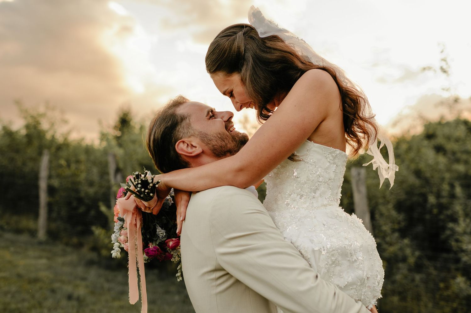 Groom lifts bride in wedding attire at sunset in a garden, sharing a romantic moment with a floral bouquet.