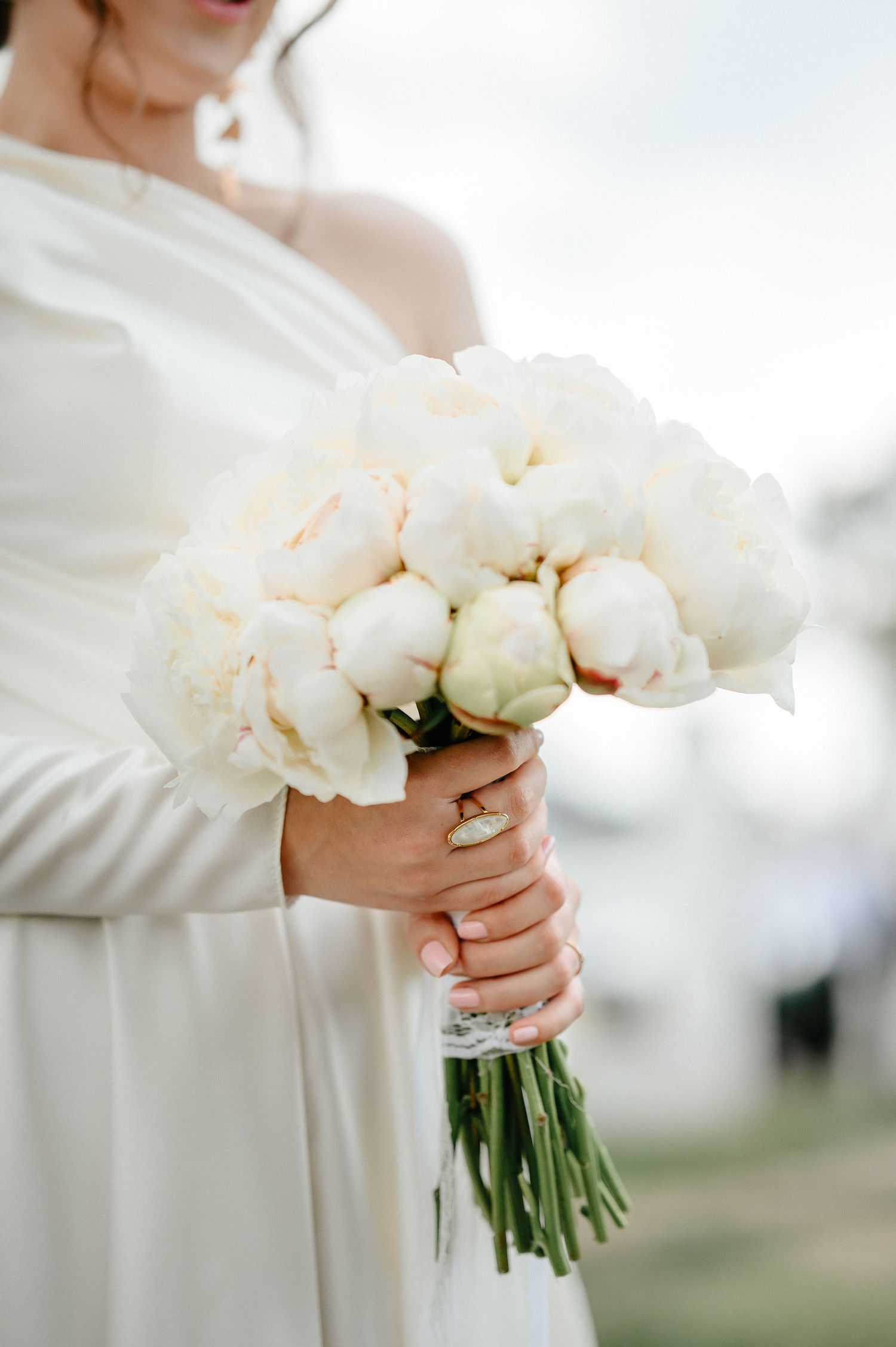 Close-up of bride holding lush white peony wedding bouquet with gold engagement ring.