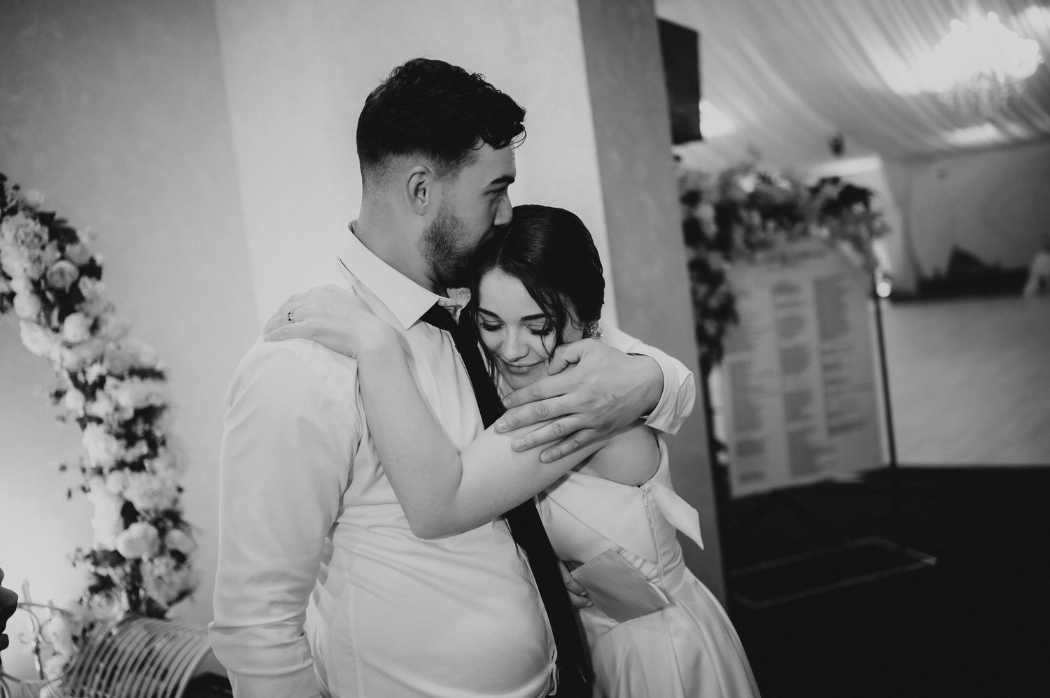 Black and white photo of groom embracing bride tenderly near floral arch at wedding ceremony.