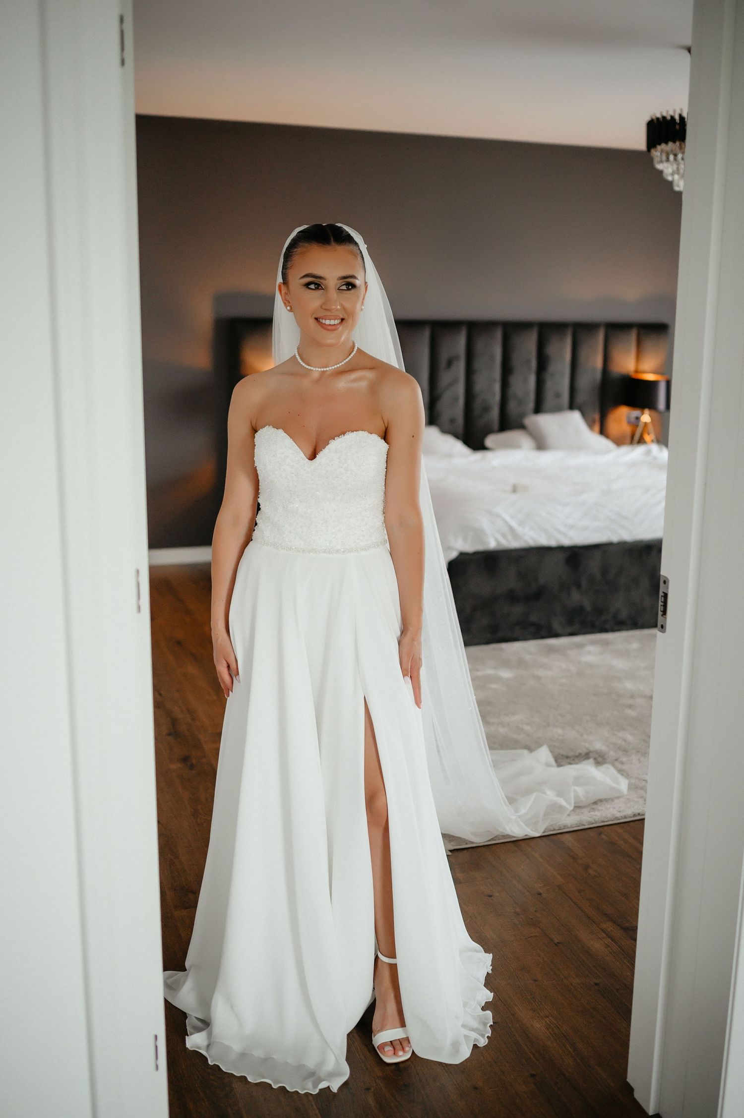 Bride in strapless white gown with veil and pearl necklace smiles in a bedroom doorway.